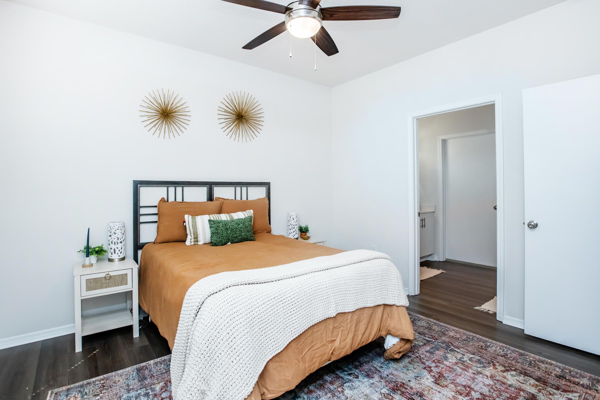 A cozy bedroom featuring a bed with an orange-brown comforter and decorative pillows. Two gold wall decor pieces are mounted above the headboard. There's a small nightstand beside the bed with a lamp and a green decorative item. A doorway leads to another room, and a colorful area rug is laid on the floor.