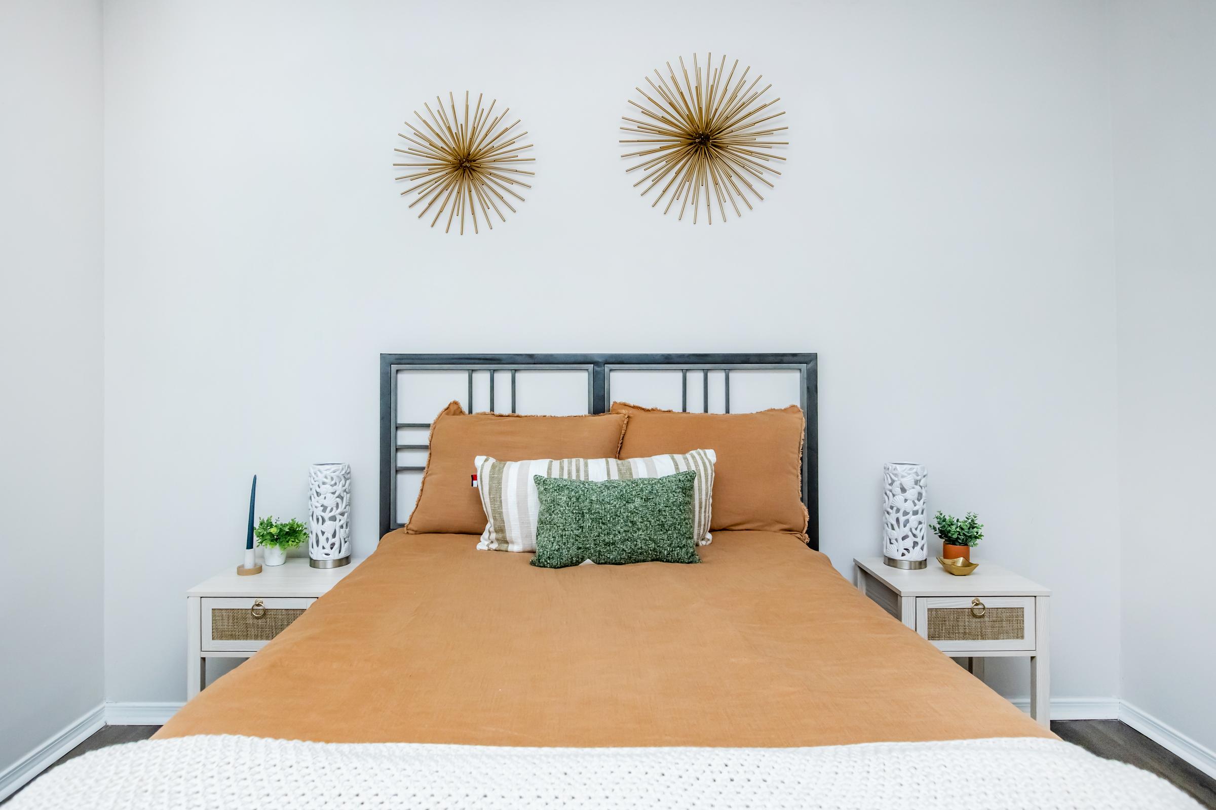 A cozy bedroom featuring a neatly made bed with a brown-orange quilt and decorative pillows. Two stylish starburst wall decorations are above the headboard. There are white bedside tables on either side, each with a small plant and a lamp, set against a clean, white wall.