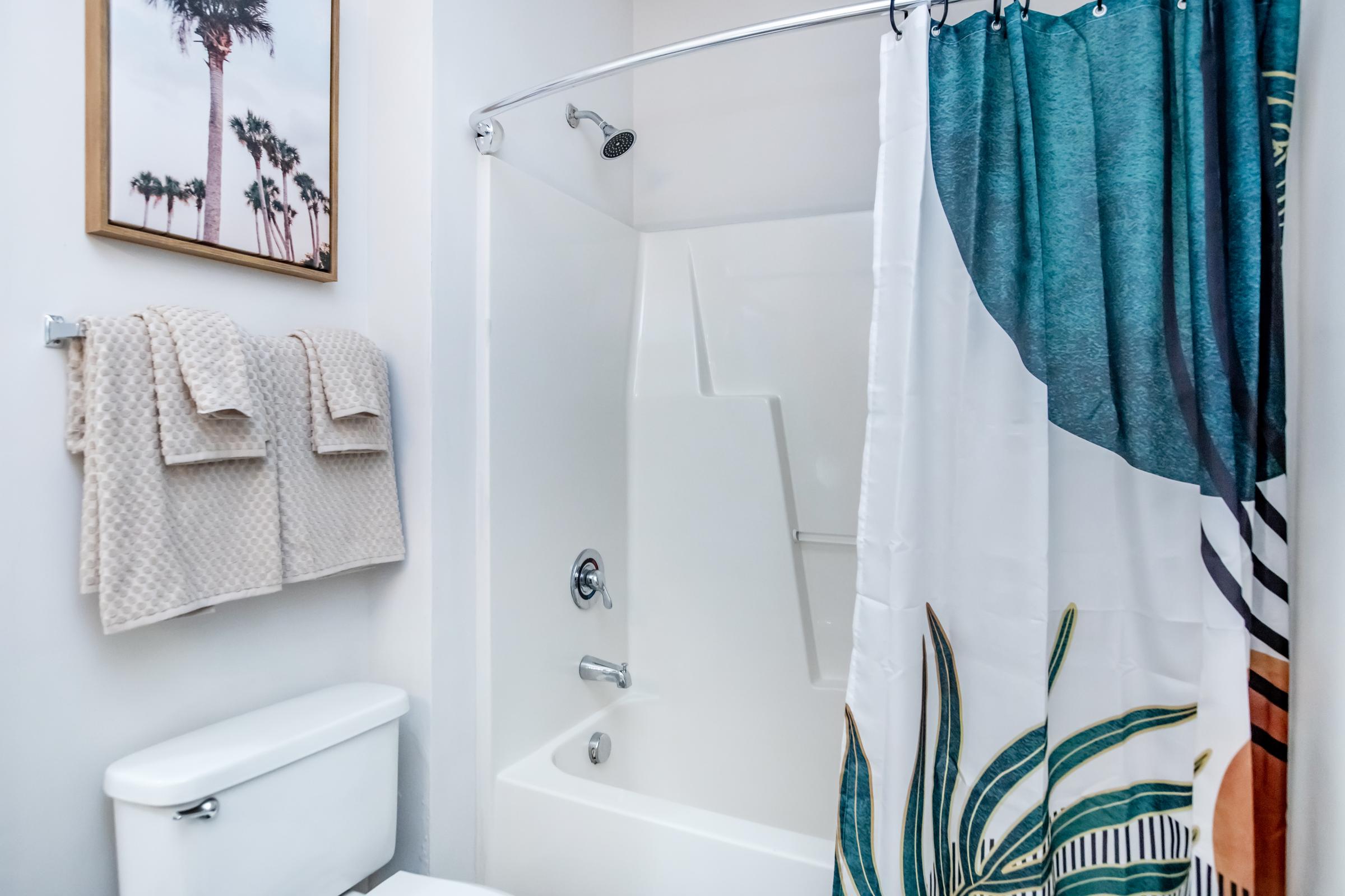A modern bathroom featuring a white shower stall with a silver showerhead, a decorative shower curtain with a tropical leaf design, neatly hung beige towels on the wall, and a white toilet. A framed picture of palm trees is visible on the wall, adding to the decor.