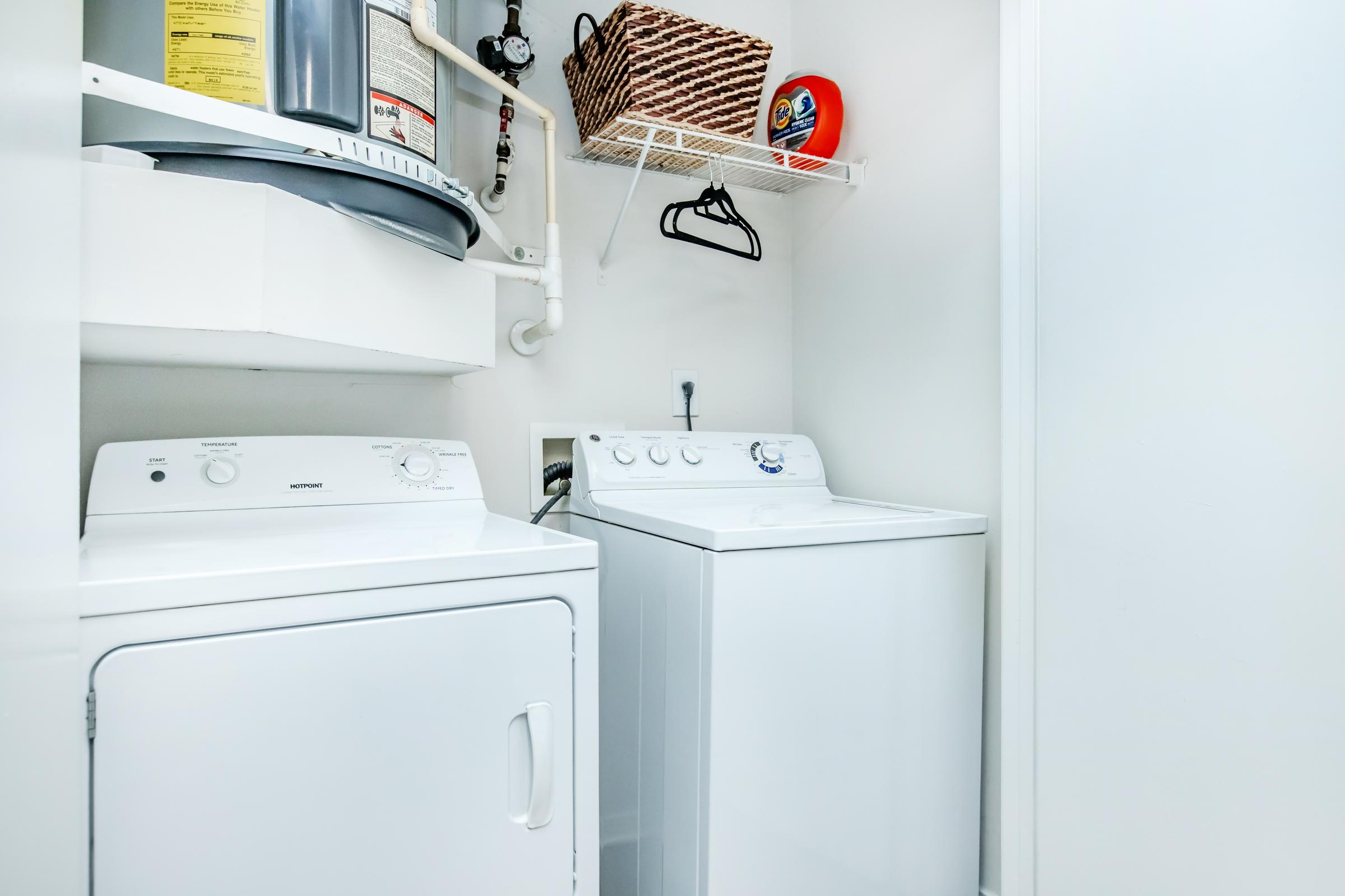 A laundry room featuring a white washer and dryer side by side. Above them is a shelf with a basket and laundry detergent. A water heater is visible on the wall, and a single hanger is attached to the shelf. The overall space is bright and organized, designed for efficient laundry tasks.