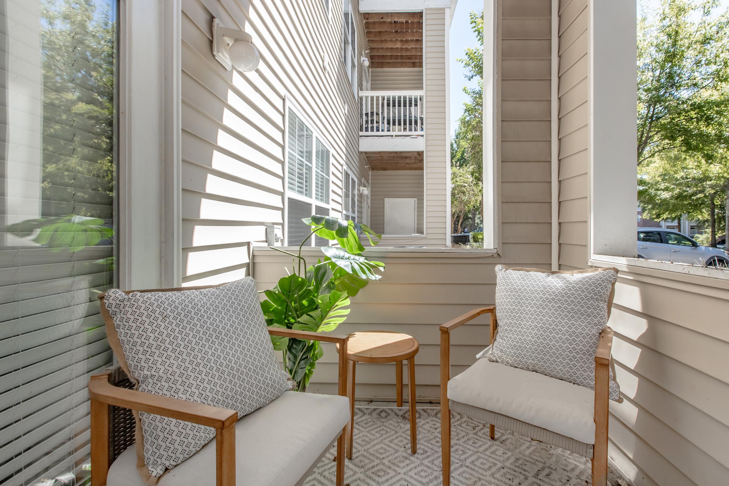 A cozy outdoor balcony featuring two wooden chairs with patterned cushions and a small round table. A green plant adds a touch of nature, and the background shows neighboring buildings and trees. Sunlight filters through, creating a warm and inviting atmosphere.