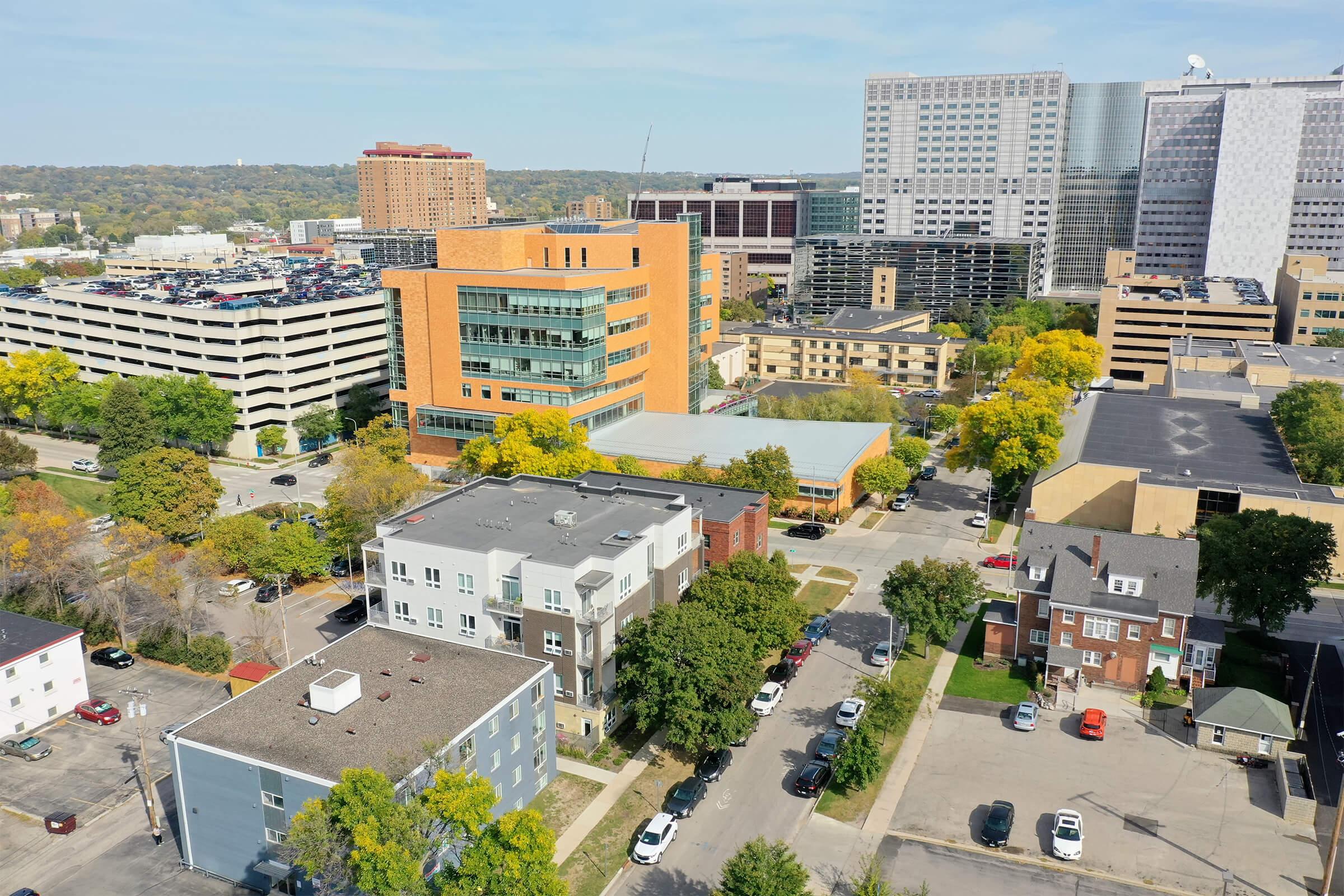 Aerial view of a cityscape featuring a mix of modern and older buildings, parking structures, and tree-lined streets in the foreground. The scene shows a vibrant urban area with various architectural styles and greenery, indicating a blend of residential and commercial spaces.