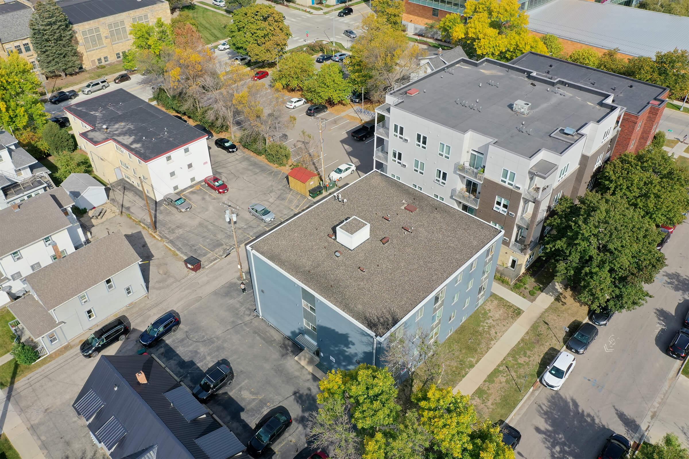 Aerial view of a residential area featuring a mix of apartment buildings, parked cars, and tree-lined streets. The image captures two multi-story buildings, one with a flat roof and the other featuring a sloped roof, surrounded by greenery and asphalt roads.
