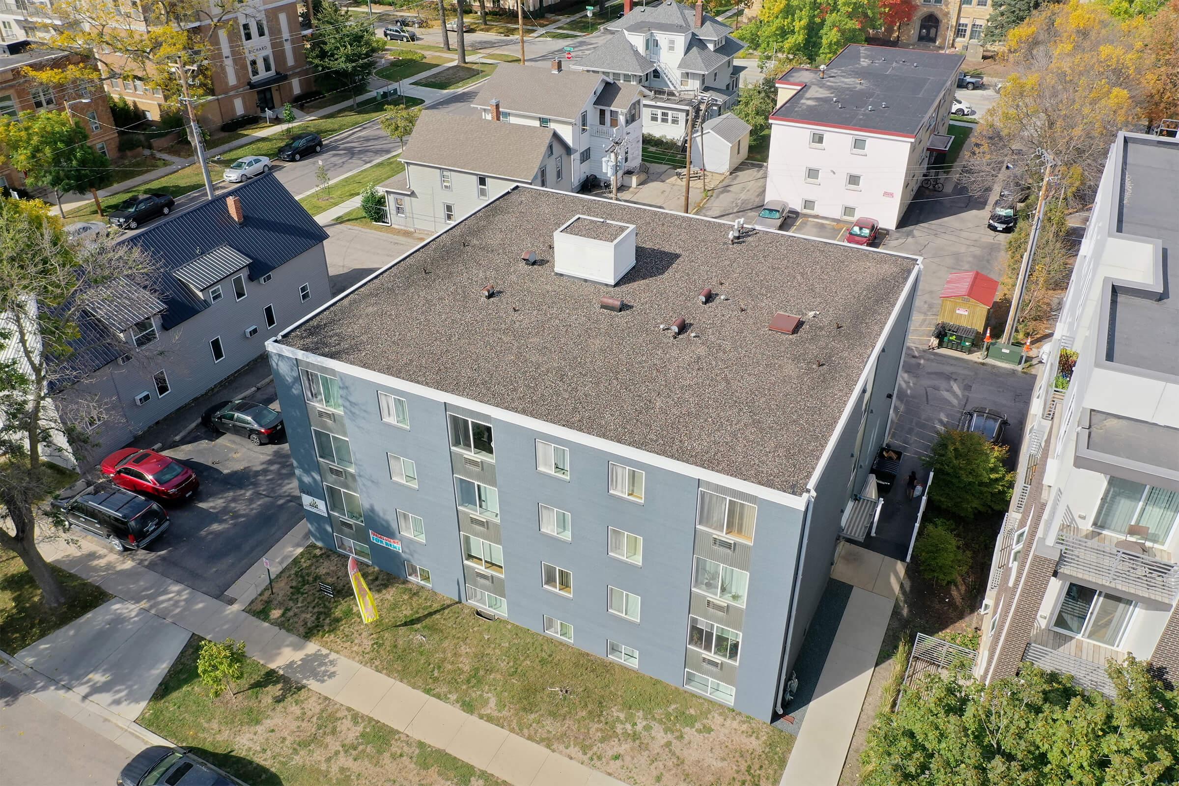 Aerial view of a mid-sized residential building with a flat roof and blue-gray exterior. Surrounding area features several other houses and buildings, with cars parked nearby. Trees with autumn foliage are visible, indicating a suburban neighborhood. The scene is clear and well-lit.