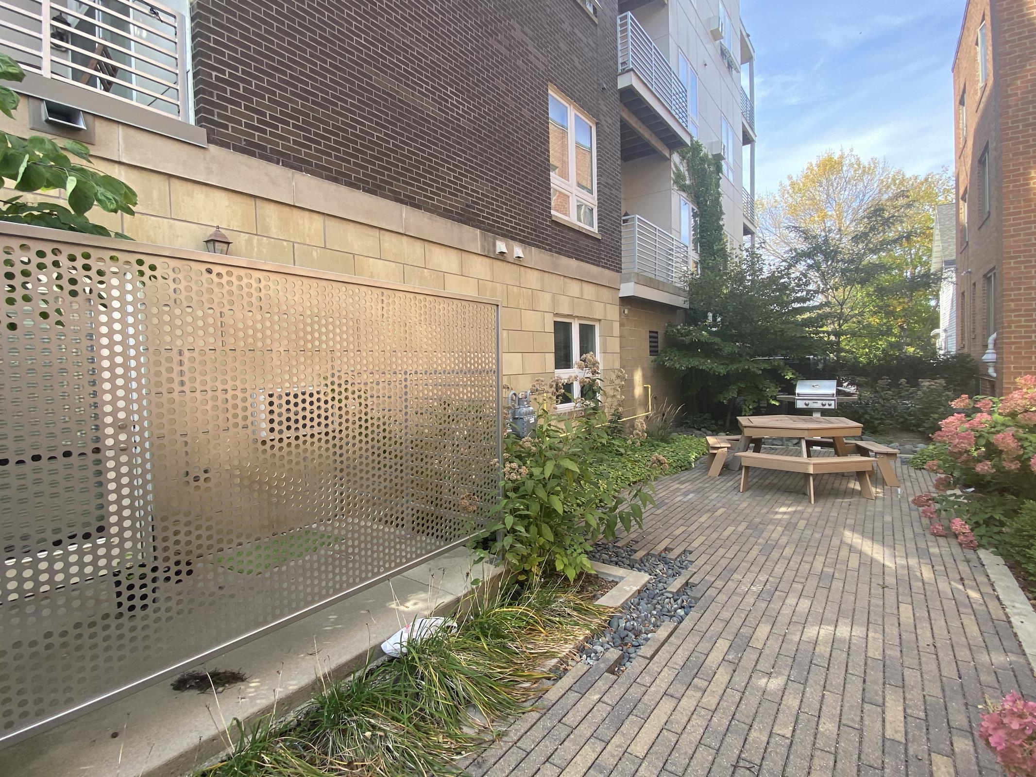 A landscaped courtyard featuring a seating area with a wooden table and benches, surrounded by greenery. A decorative metal panel with circular cutouts is positioned on the left, with a brick pathway leading through the space. Nearby, there's a grill and trees in the background under a clear sky.