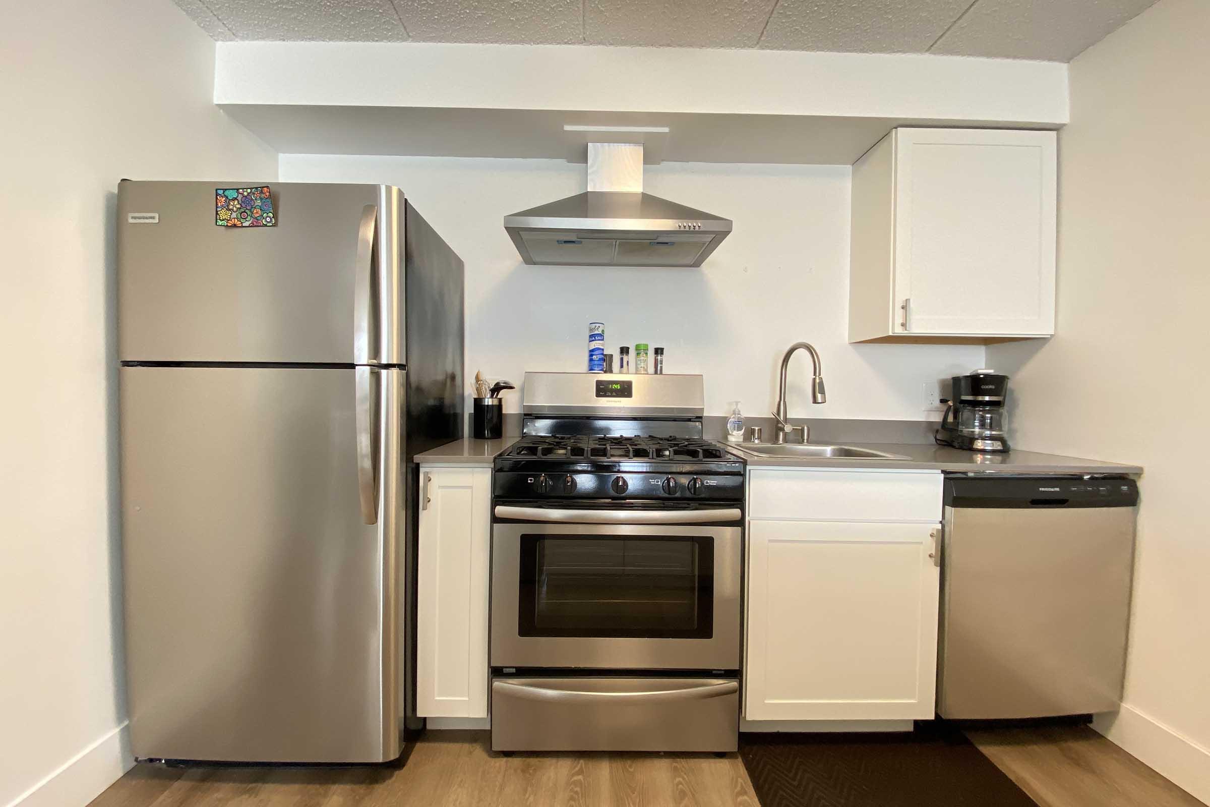 A modern kitchen featuring stainless steel appliances including a refrigerator, oven, and dishwasher. There is a range hood above the stove, white cabinetry, a sink, and a coffee maker on the counter. The floor is wooden, and the walls are painted white.