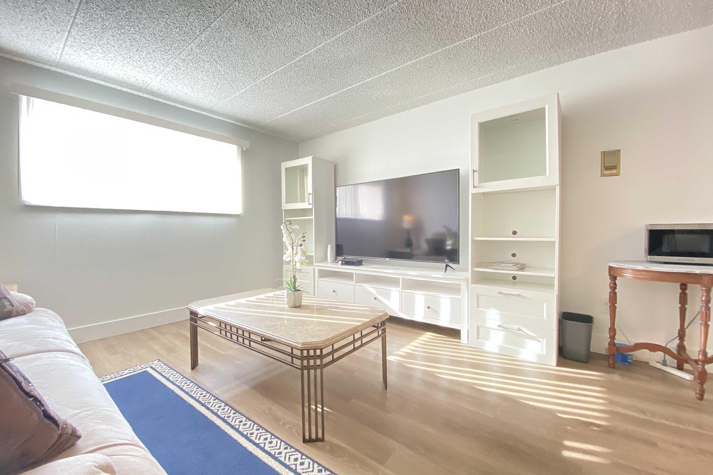 A bright and airy living room featuring a white sofa, a large flat-screen TV on a white media console, and a glass coffee table with a decorative vase. There is a small side table and a microwave in the background, with natural light streaming in from a window, enhancing the inviting atmosphere.