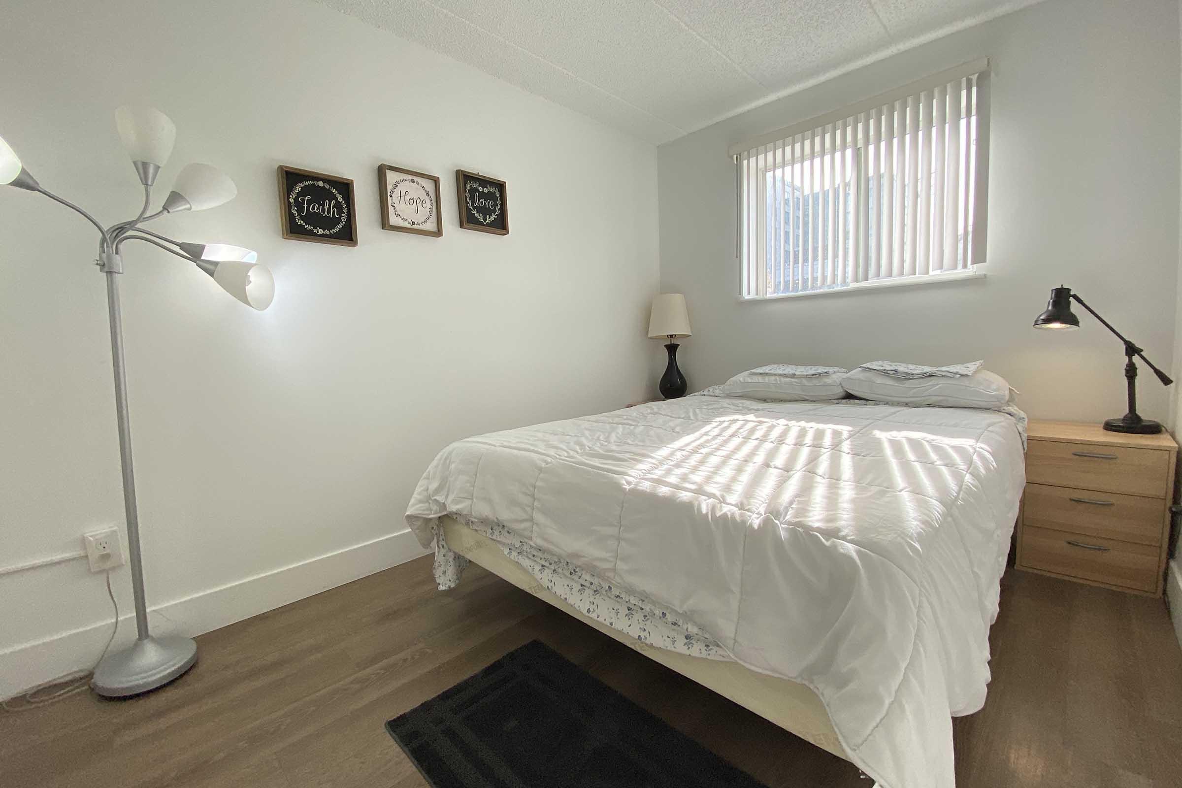 A cozy bedroom featuring a queen-sized bed with white bedding, a nightstand with a lamp, and a decorative floor lamp. The walls are adorned with four framed signs. A window with horizontal blinds allows natural light to illuminate the room, creating a warm and inviting atmosphere.