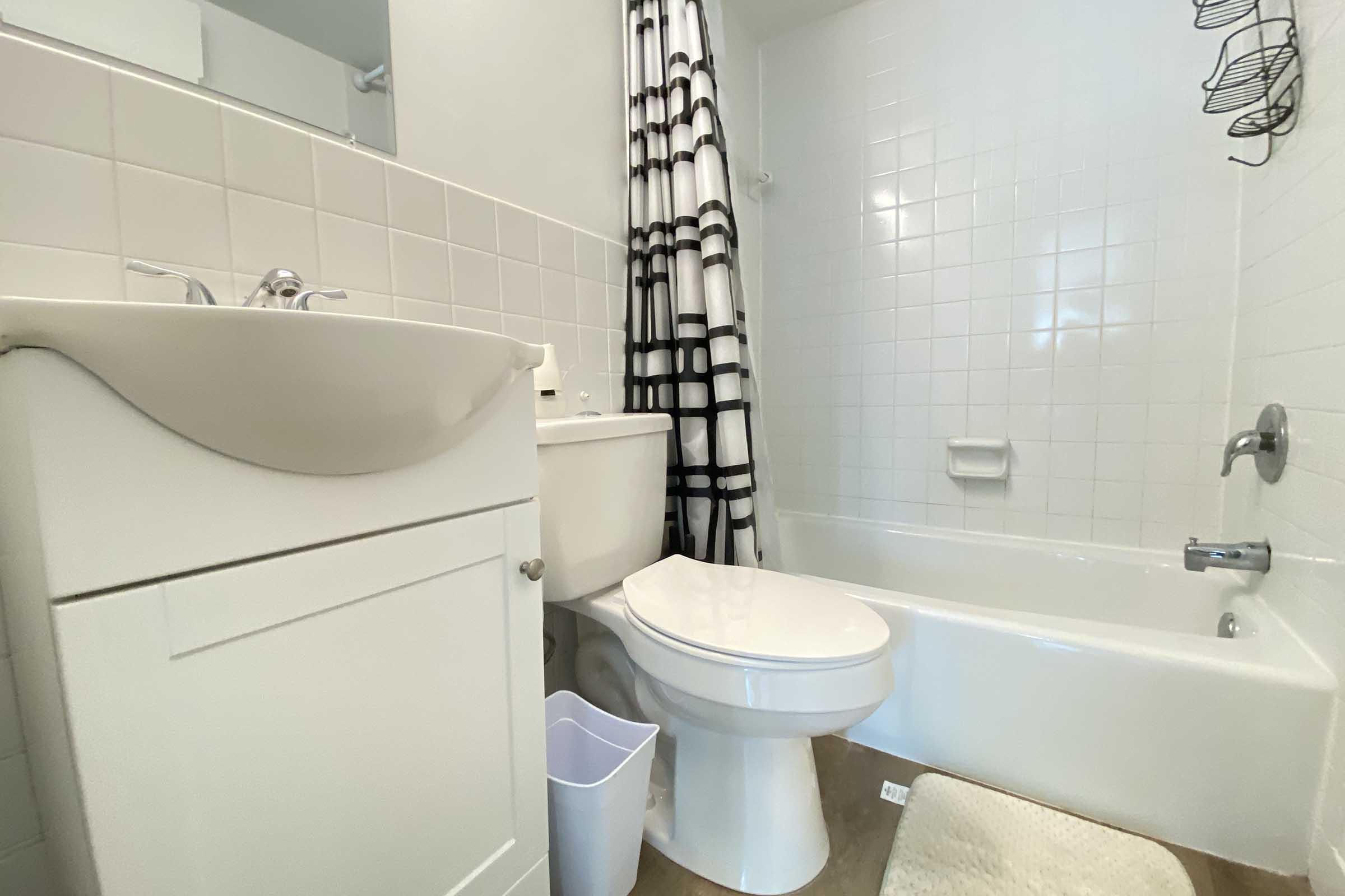 A clean bathroom featuring a white bathtub, a toilet, and a sink. There is a black and white striped shower curtain, a small trash bin, and a woven bath mat on the floor. The walls are tiled in light colors, creating a bright and airy atmosphere.