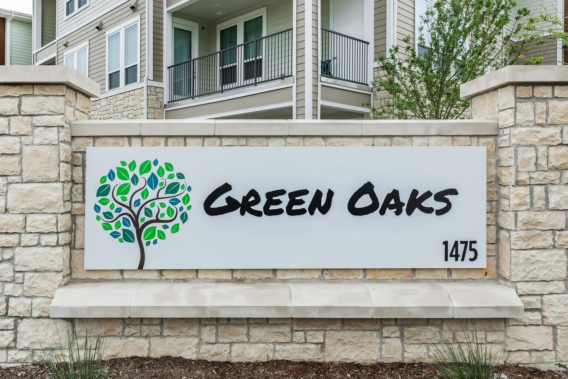 Sign for Green Oaks located at 1475, featuring a stylized tree graphic with green leaves. The background is a light stone wall, emphasizing the text "Green Oaks" in bold, black font. The setting suggests a residential community.