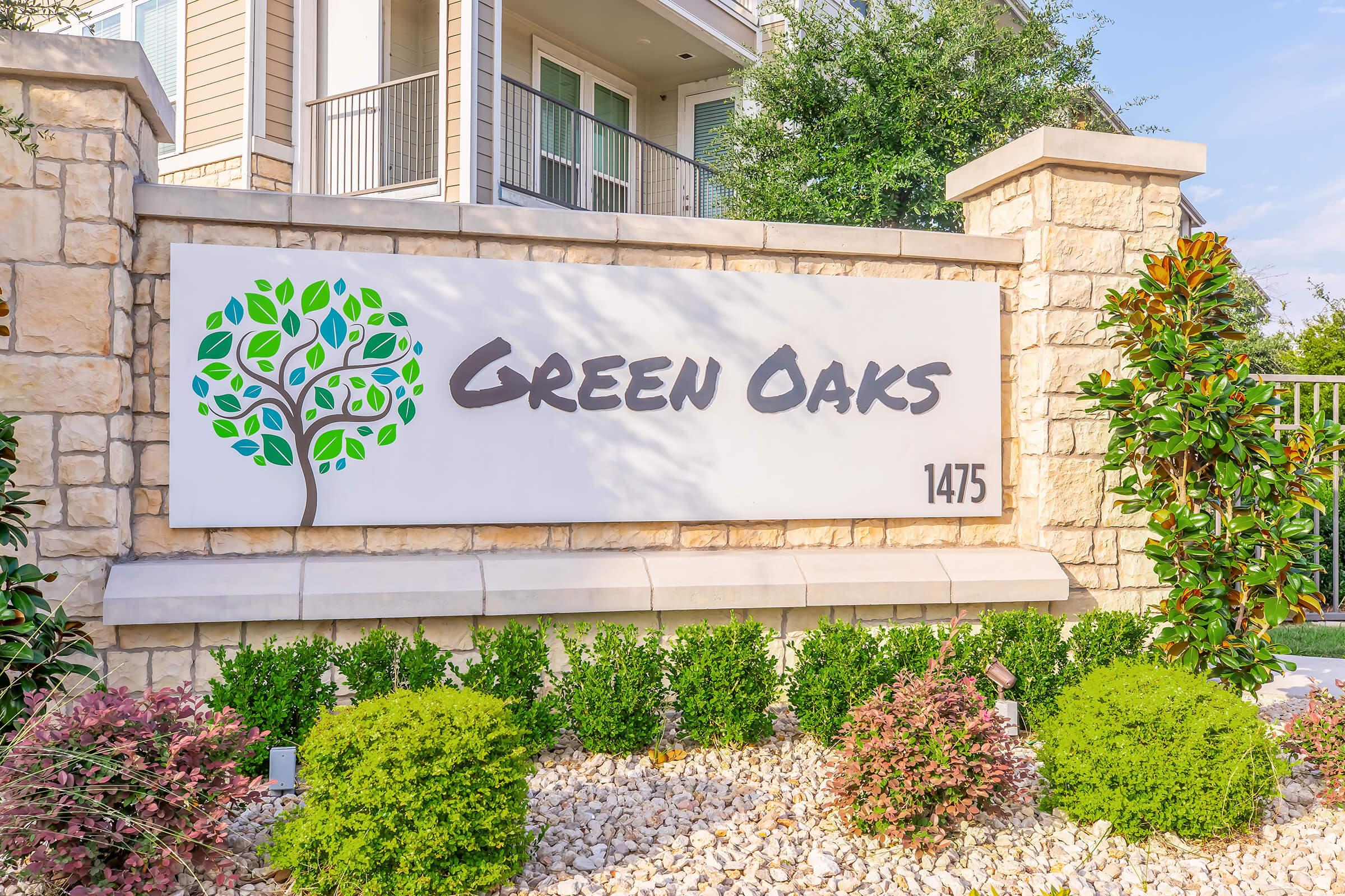 Sign for "Green Oaks" featuring a stylized tree logo. The sign is made of stone and is surrounded by neatly trimmed shrubbery and landscaping. The background shows a multi-story building and green trees, indicating a well-kept residential area. Address "1475" is also displayed on the sign.