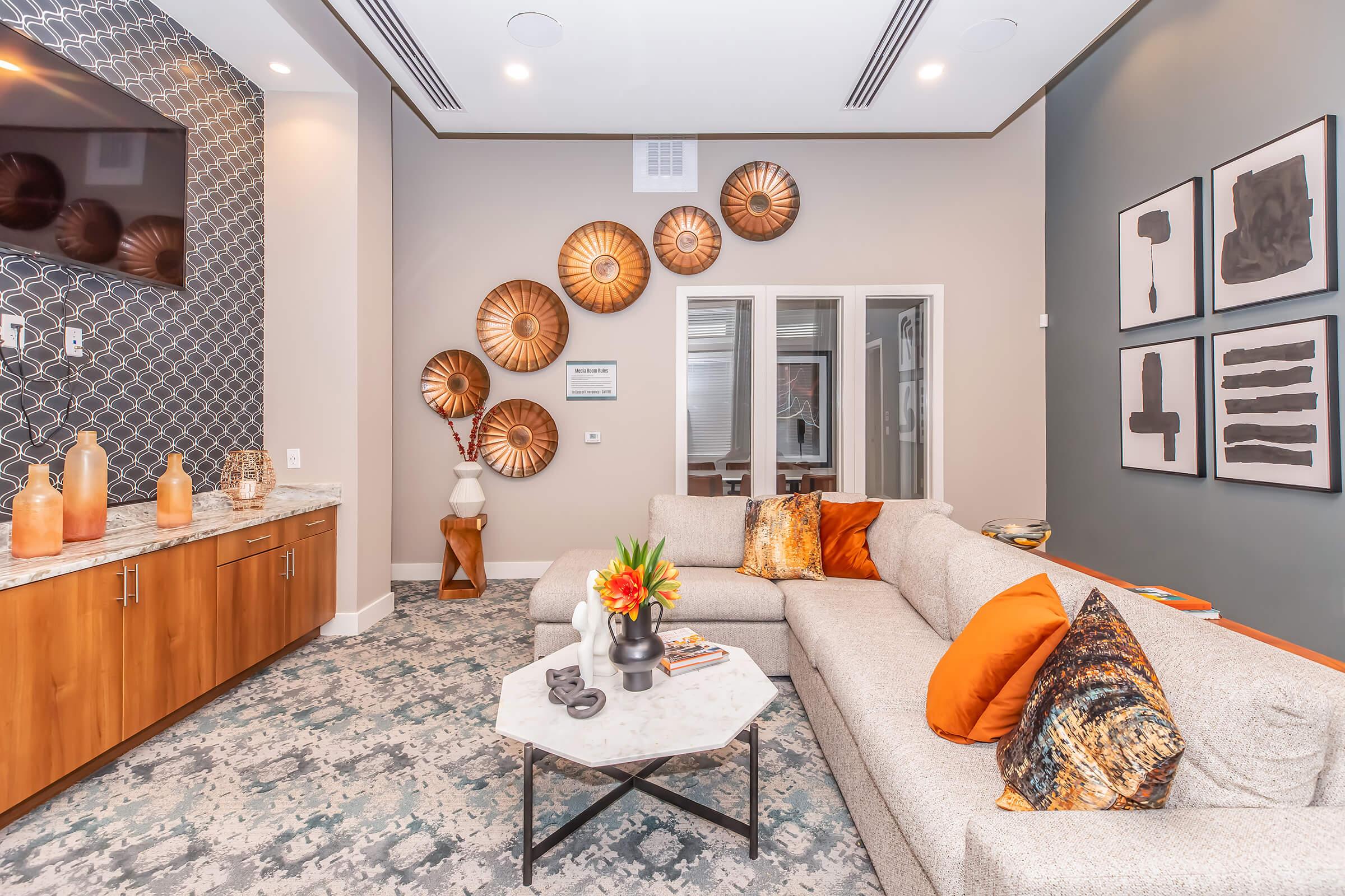 A modern living room featuring a light-colored sectional sofa adorned with bright orange and patterned throw pillows. A small coffee table sits in front, topped with a vase of flowers. The walls display decorative round wall art and framed abstract prints. A TV is mounted above a stylish cabinet.