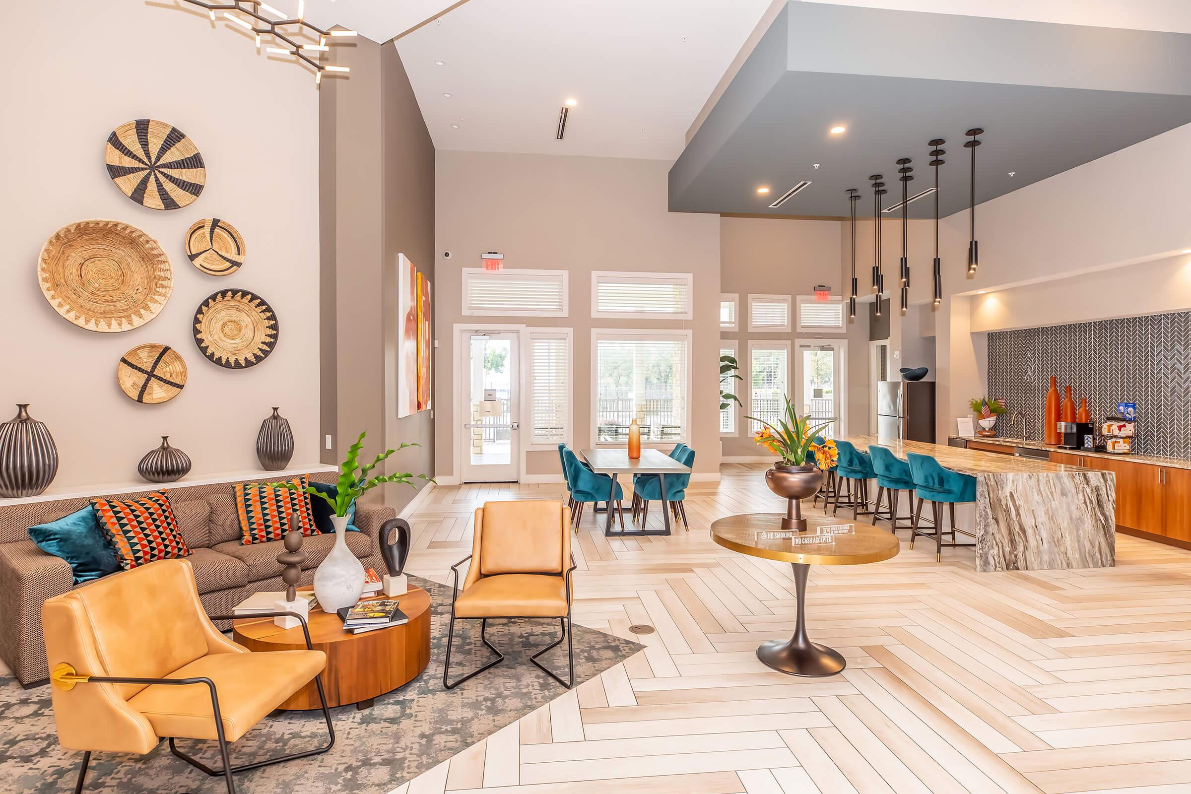 A modern lounge area featuring a cozy seating arrangement with a brown sofa and stylish armchairs. Decorative wall plates adorn one wall, while a large table and chairs are set nearby. Bright natural light floods through large windows, and a sleek bar area with decorative elements adds to the inviting atmosphere.