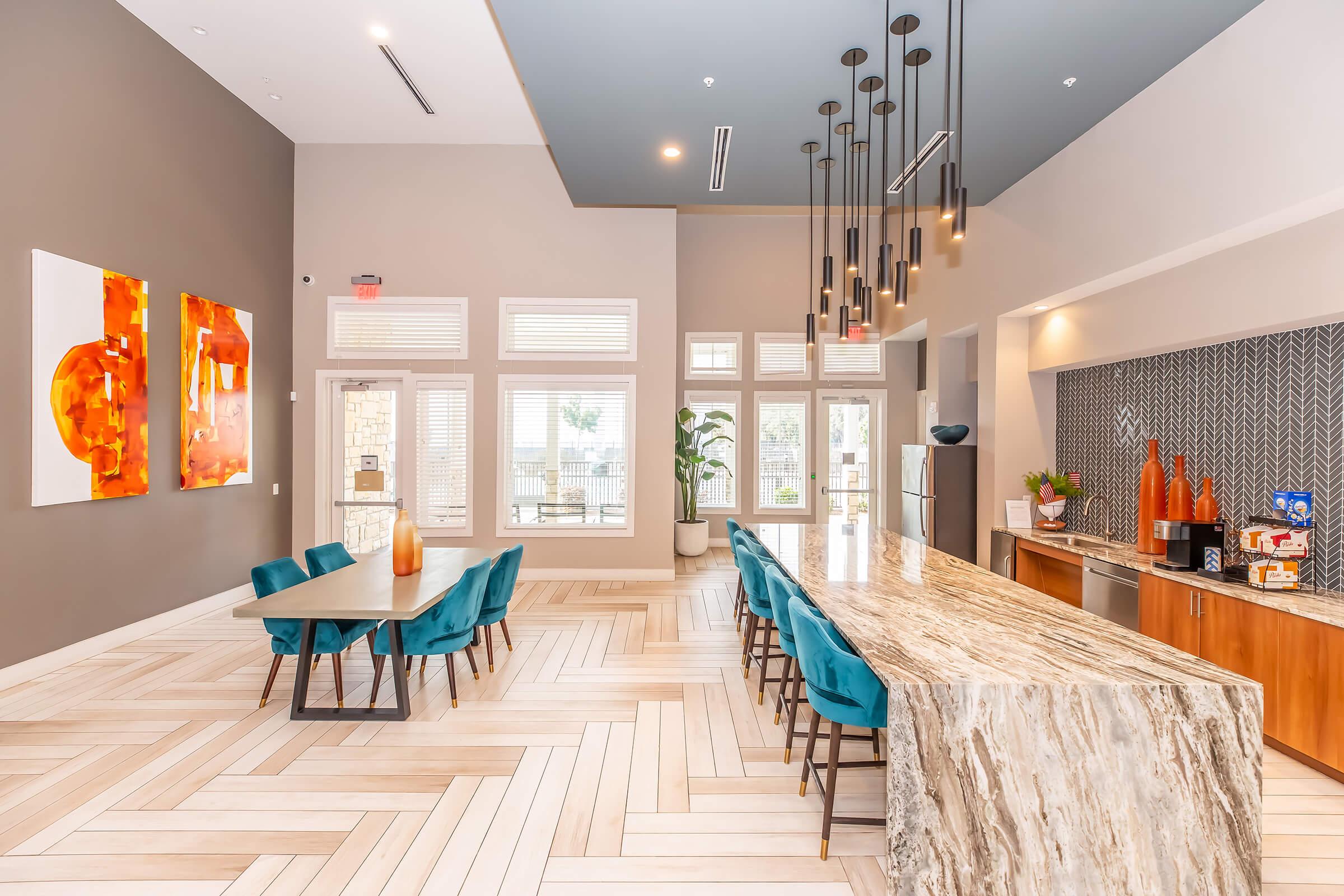 A modern, spacious community kitchen and dining area featuring a large marble countertop, a sleek dining table with turquoise chairs, and hanging light fixtures. The walls are painted in neutral tones, accented with vibrant artwork, and large windows provide natural light. A kitchenette is visible with appliances and decor.