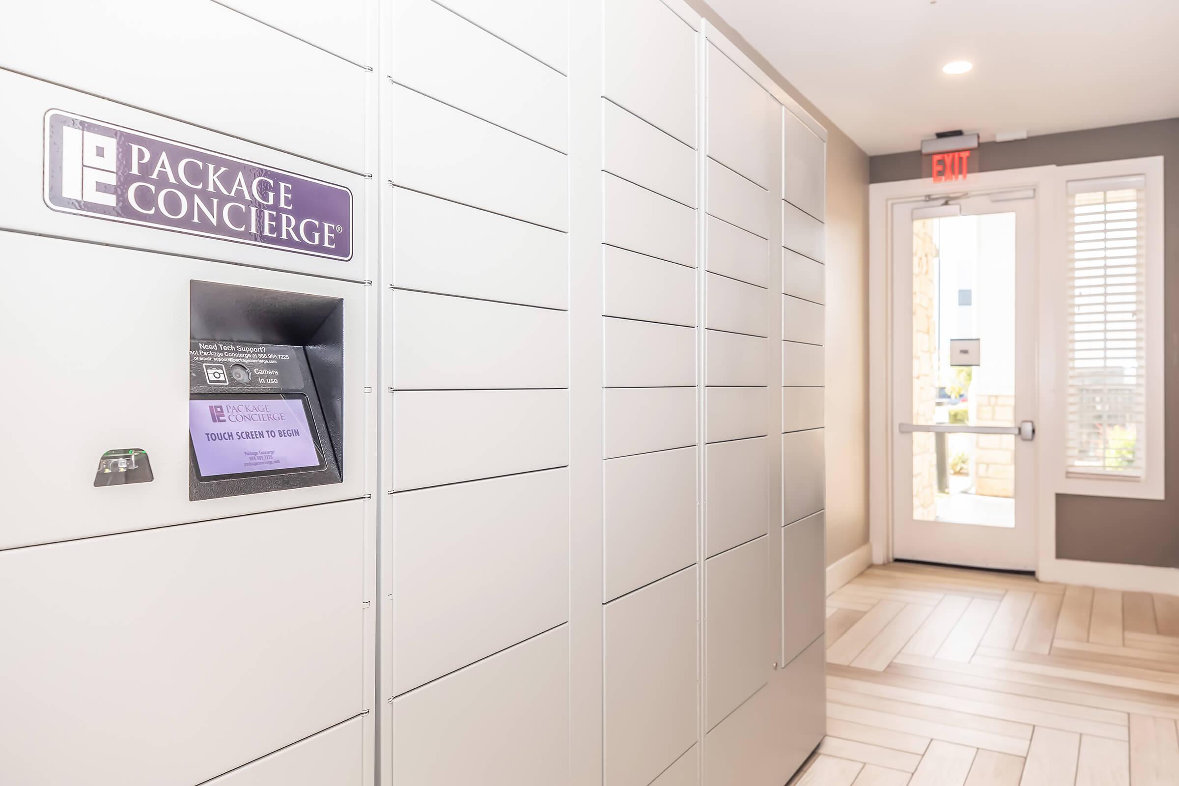 A modern package concierge area featuring gray lockers for package delivery. A digital screen displays instructions for package retrieval. The entrance door is visible in the background, leading to an outdoor area with natural light.