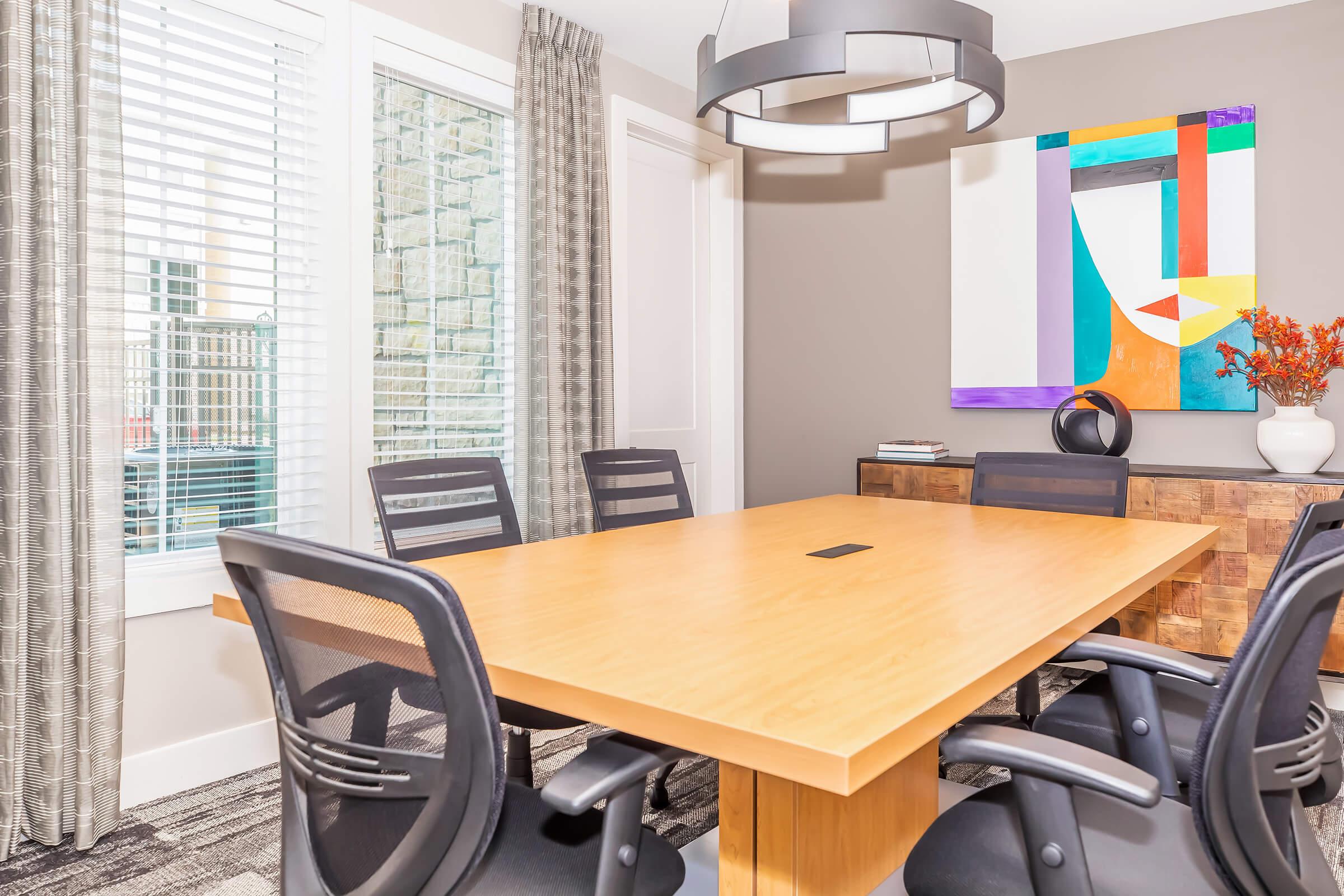 A modern conference room featuring a large wooden table surrounded by black office chairs. Natural light streams in through window blinds, and a colorful abstract painting hangs on the wall. A decorative vase with flowers adds a touch of color, while a stylish pendant light hangs overhead.