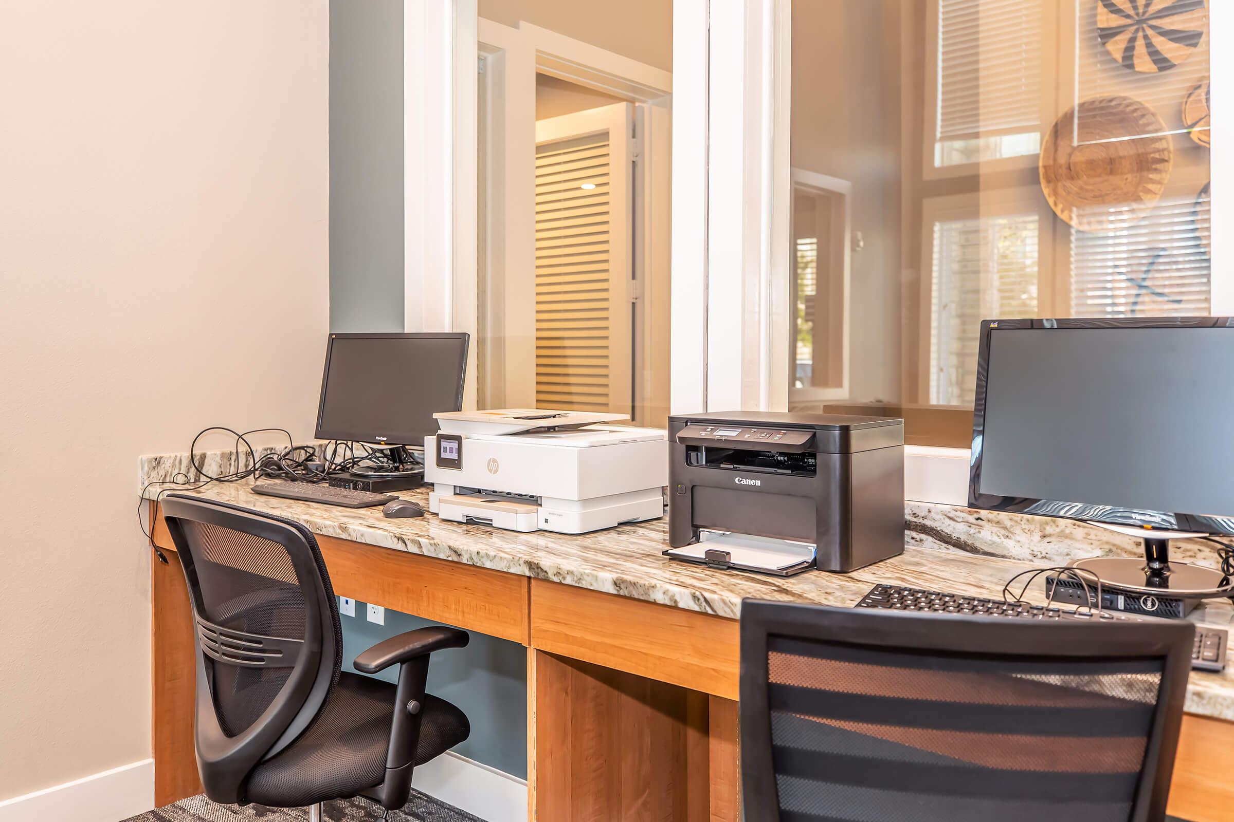 Office workspace featuring a wooden desk with two computer monitors, a printer, and a scanner. The space has a light-colored wall, a granite countertop, and two office chairs. Natural light enters through a nearby window, enhancing the modern and organized atmosphere.