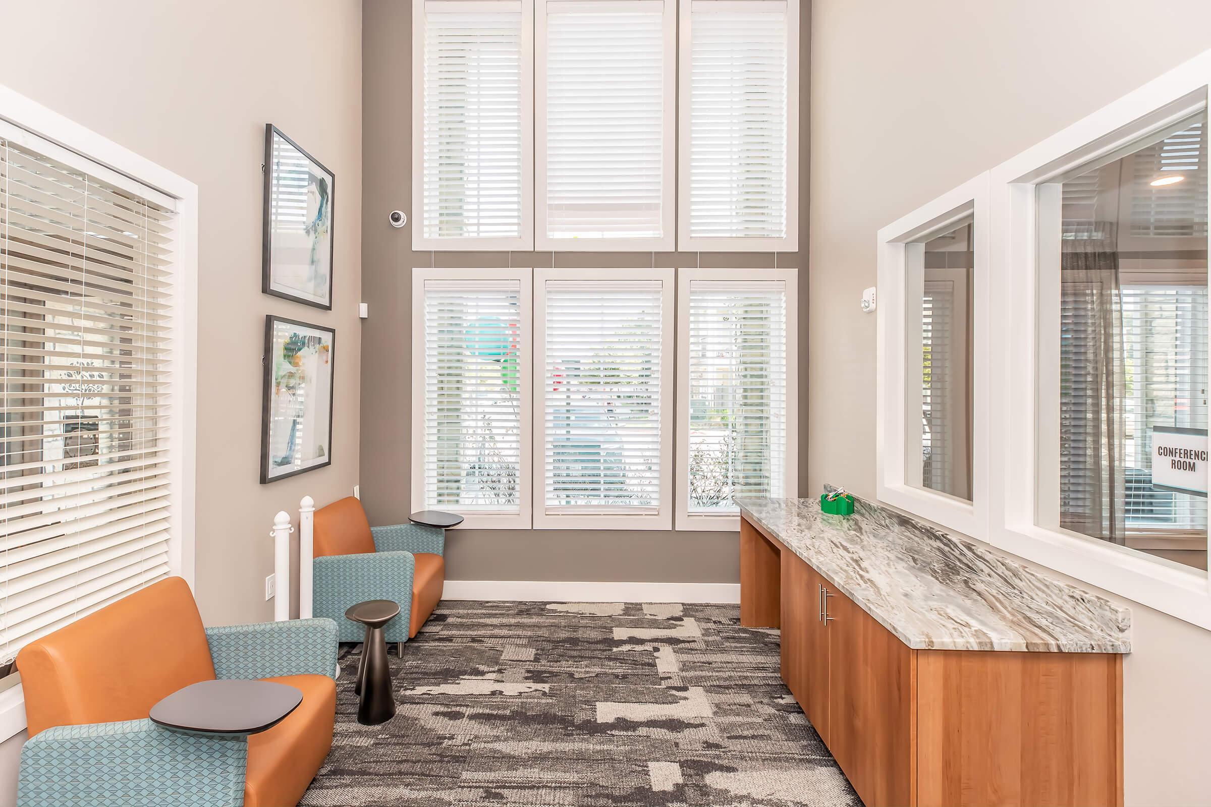 Interior of a modern lobby featuring large windows with light-colored blinds, two blue chairs and brown accent chairs, a sleek granite countertop along the wall, and abstract artwork displayed. The space has a clean, inviting atmosphere with a neutral color palette and contemporary design.