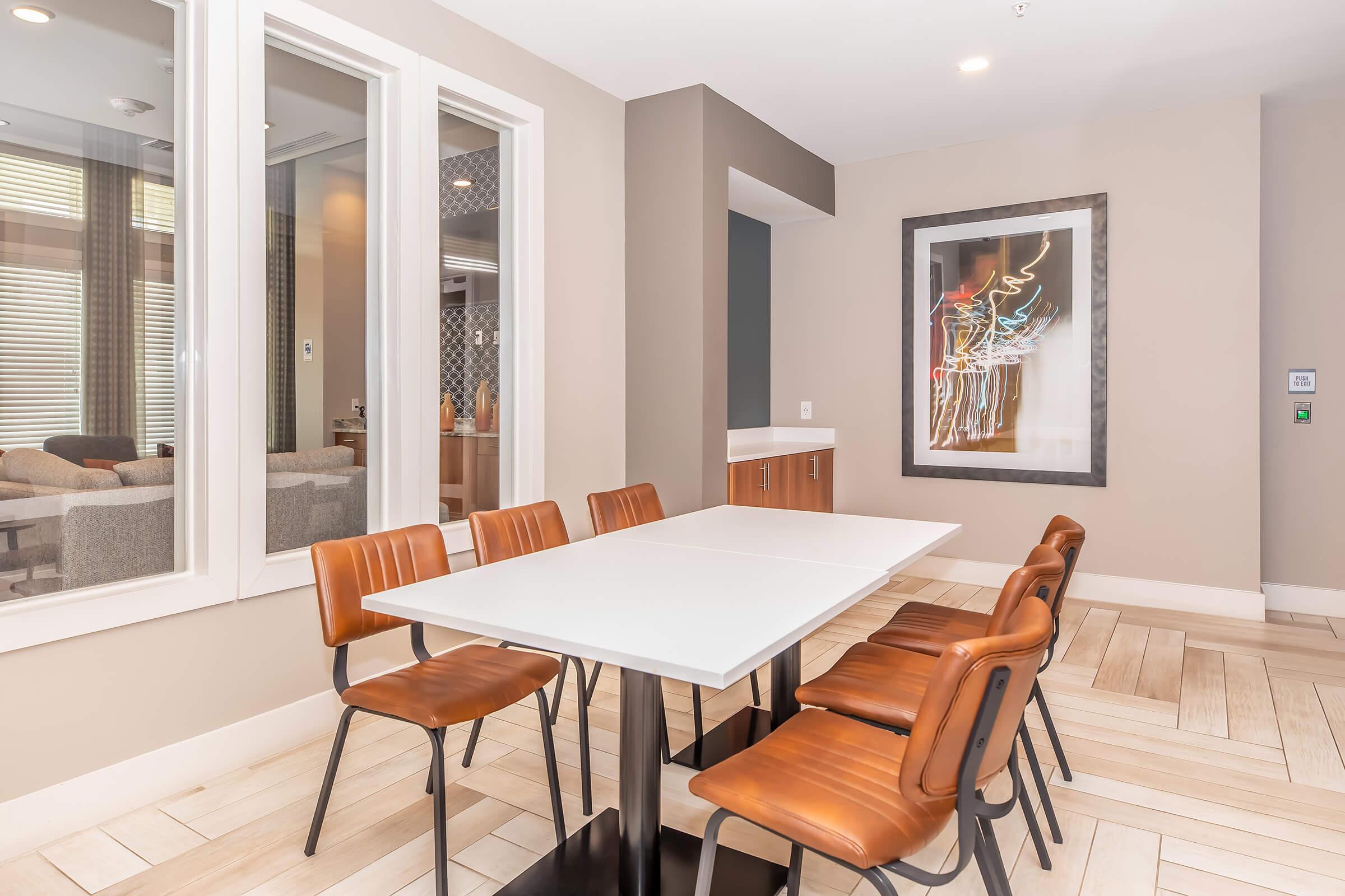 A modern dining area featuring a white table surrounded by eight brown leather chairs. The walls are painted in neutral tones, with a large abstract artwork on one side. Large windows provide natural light, and a cozy living space is visible through a glass partition in the background.