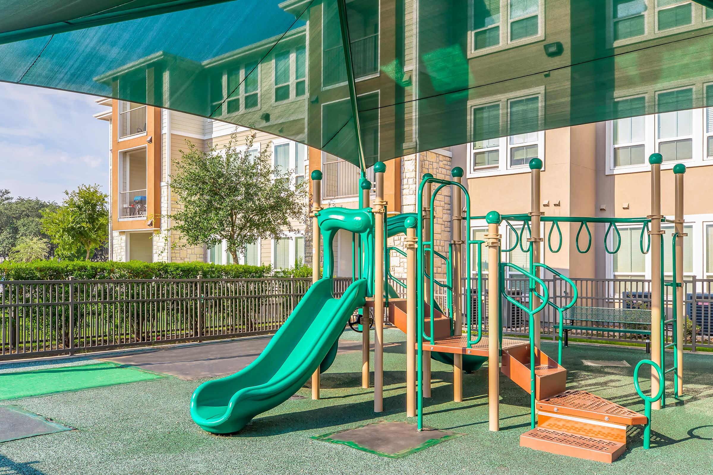 A green playground featuring a slide and climbing structure, shaded by a canopy. The area is surrounded by a fence and overlooks an apartment complex with trees and landscaping in the background. The ground is covered with safety padding.