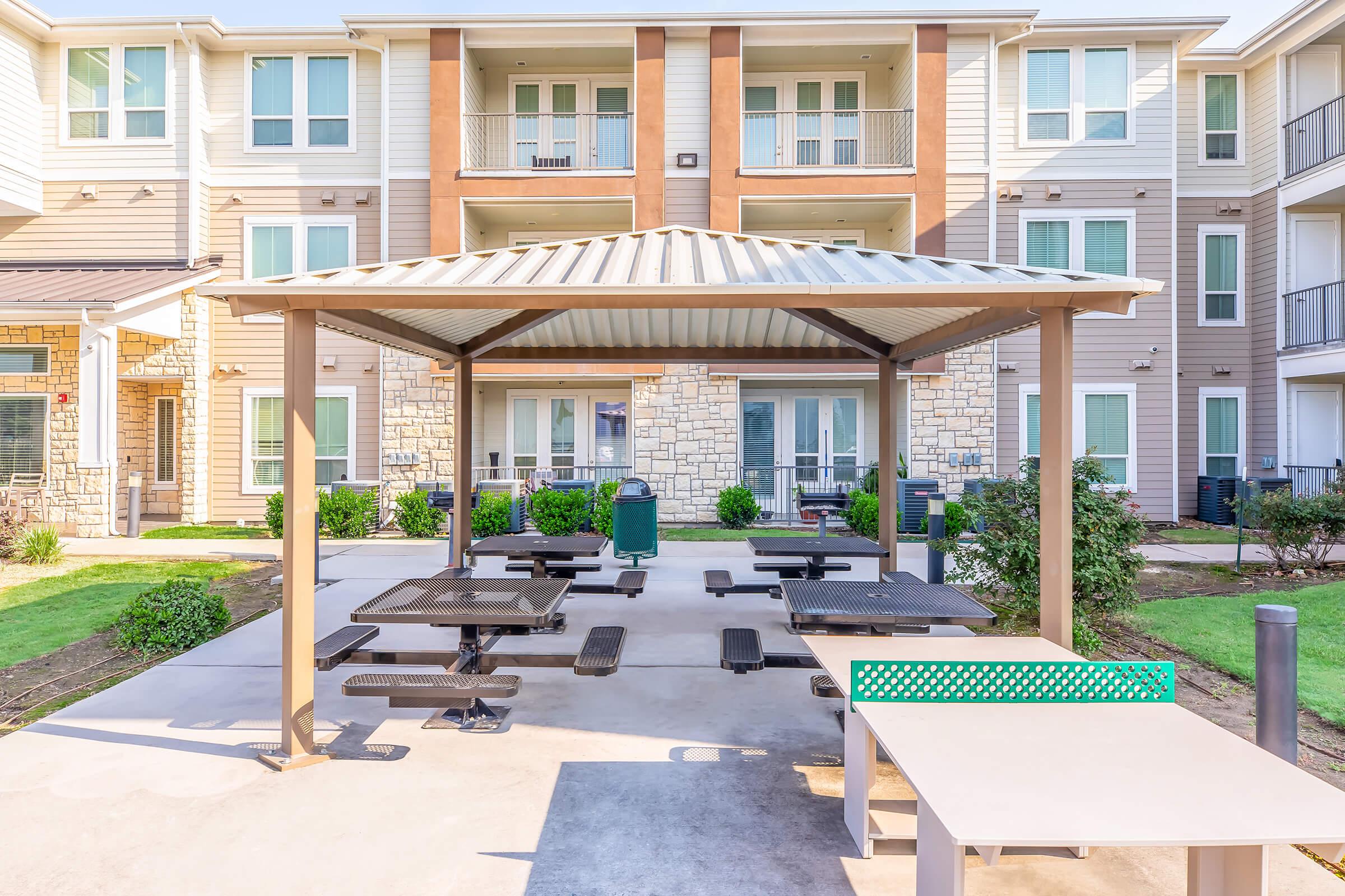 Covered outdoor seating area with picnic tables, surrounded by green grass and shrubs, adjacent to a modern apartment building with multiple balconies and large windows. The area is well-lit and inviting, ideal for social gatherings or relaxation.