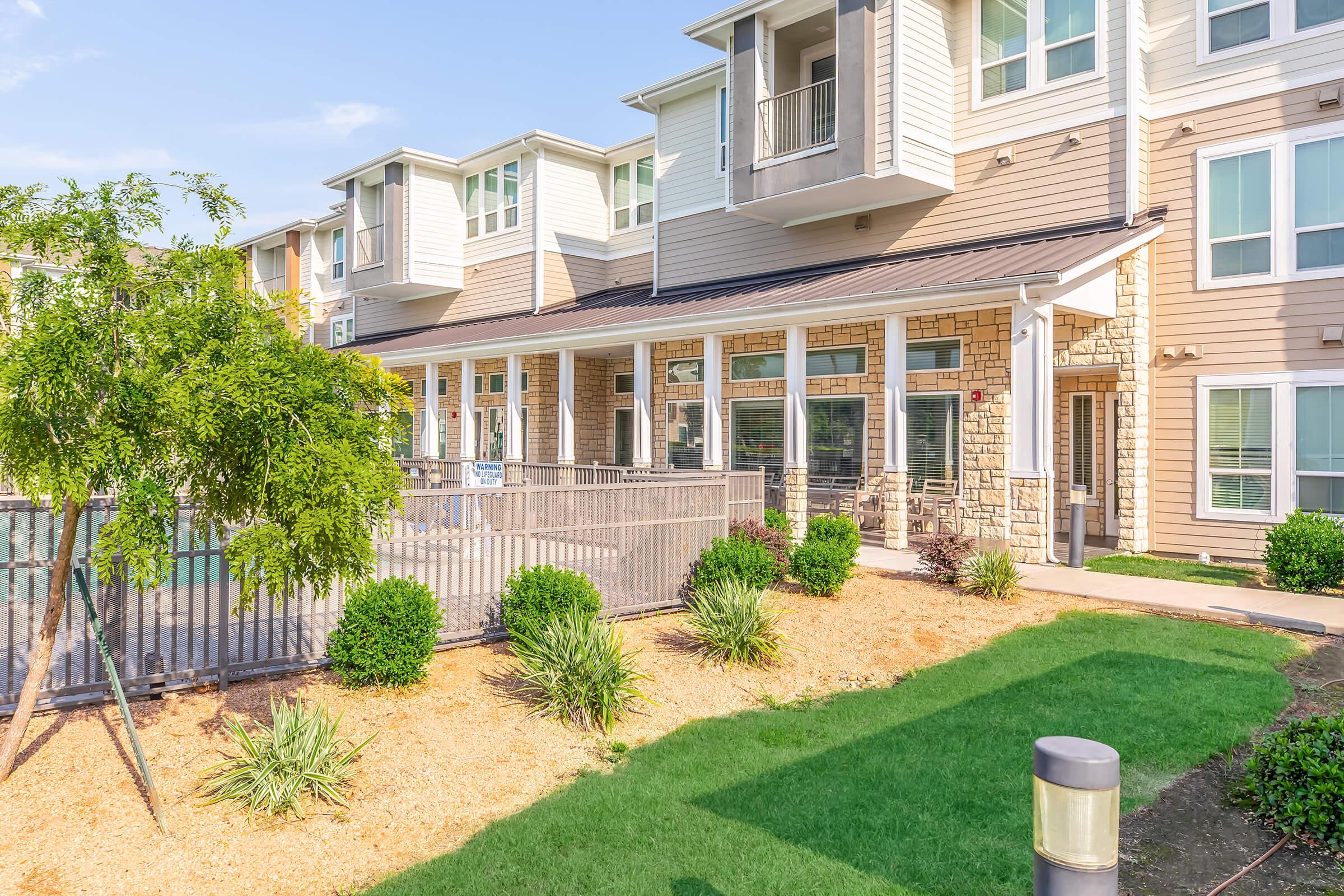 A landscaped area in front of a modern apartment building featuring multiple stories, balconies, and large windows. The yard includes manicured grass, small bushes, and a fence, creating an inviting outdoor space.