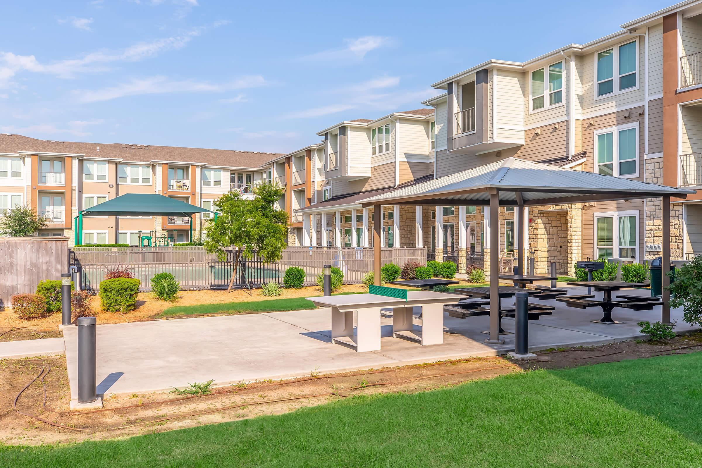 A well-maintained outdoor area featuring a concrete patio with picnic tables and a grill, surrounded by landscaped grass and small trees. In the background, there are multi-story residential buildings, and a shaded gazebo provides additional seating. The sky is clear and sunny.