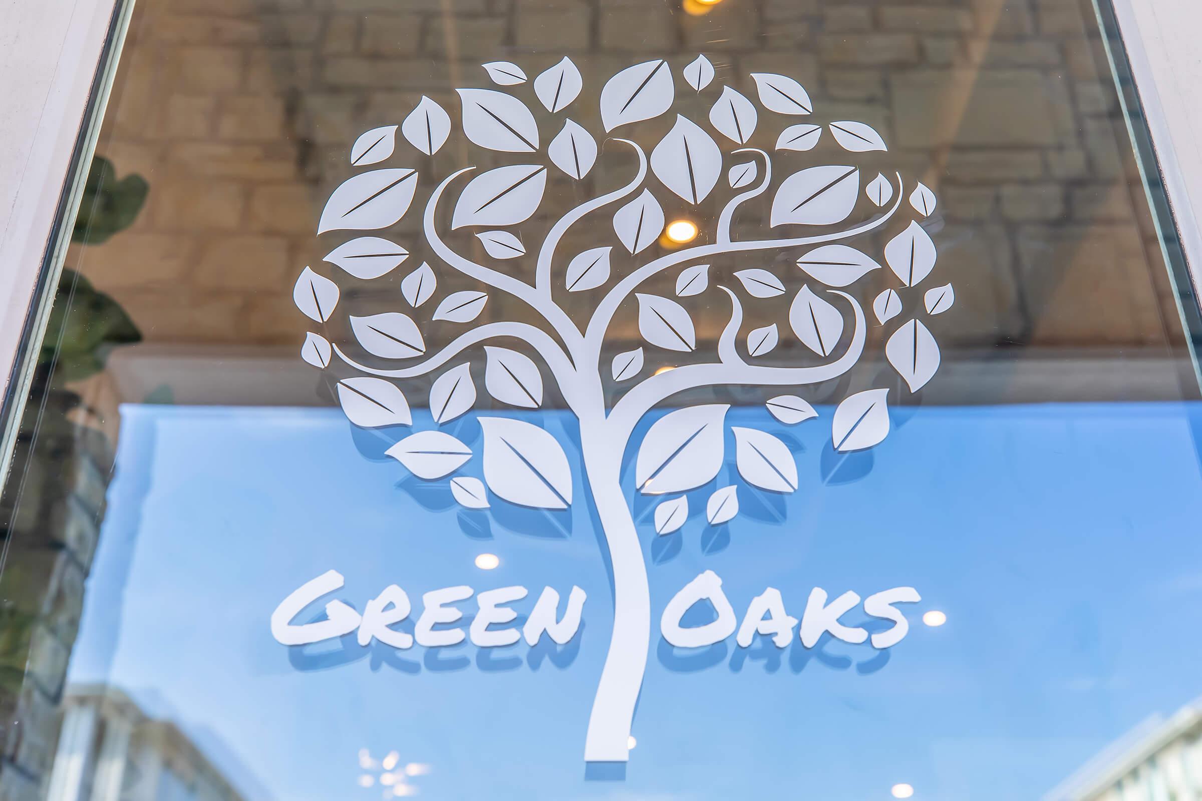 A modern, stylized tree with numerous leaves, serving as a logo, is prominently displayed on a glass surface. Below the tree, the words "GREEN OAKS" are written in bold, clear letters. The background reflects a light-colored stone wall, enhancing the design.
