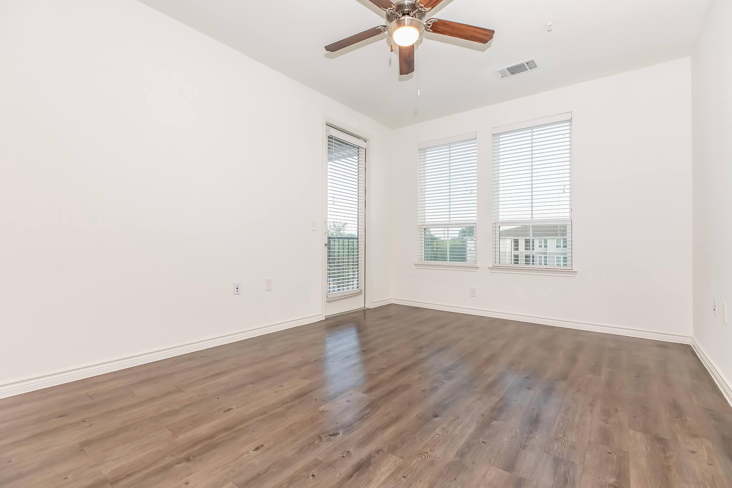 Empty room with light wood laminate flooring, a ceiling fan with wooden blades, and three windows with blinds. One window leads to a balcony, with a view of the outdoors visible through the glass door. Walls are painted white, creating a bright and airy atmosphere.