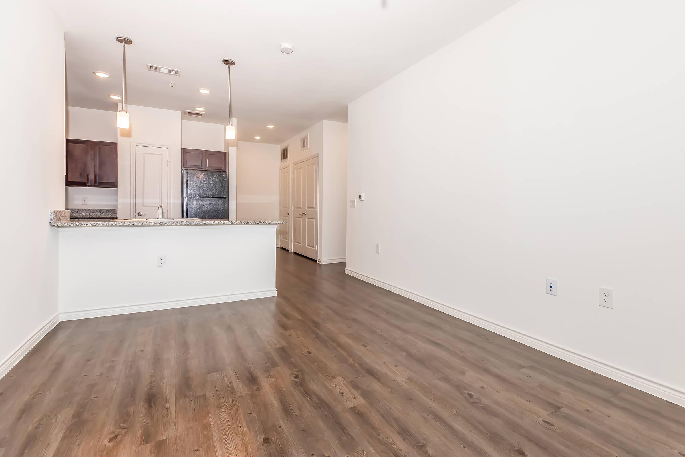 A modern, empty living space featuring light wood flooring, white walls, and a large window. The kitchen area includes a countertop with bar seating, dark cabinets, and appliances visible in the background. The layout emphasizes an open concept design, creating a spacious feel in the room.