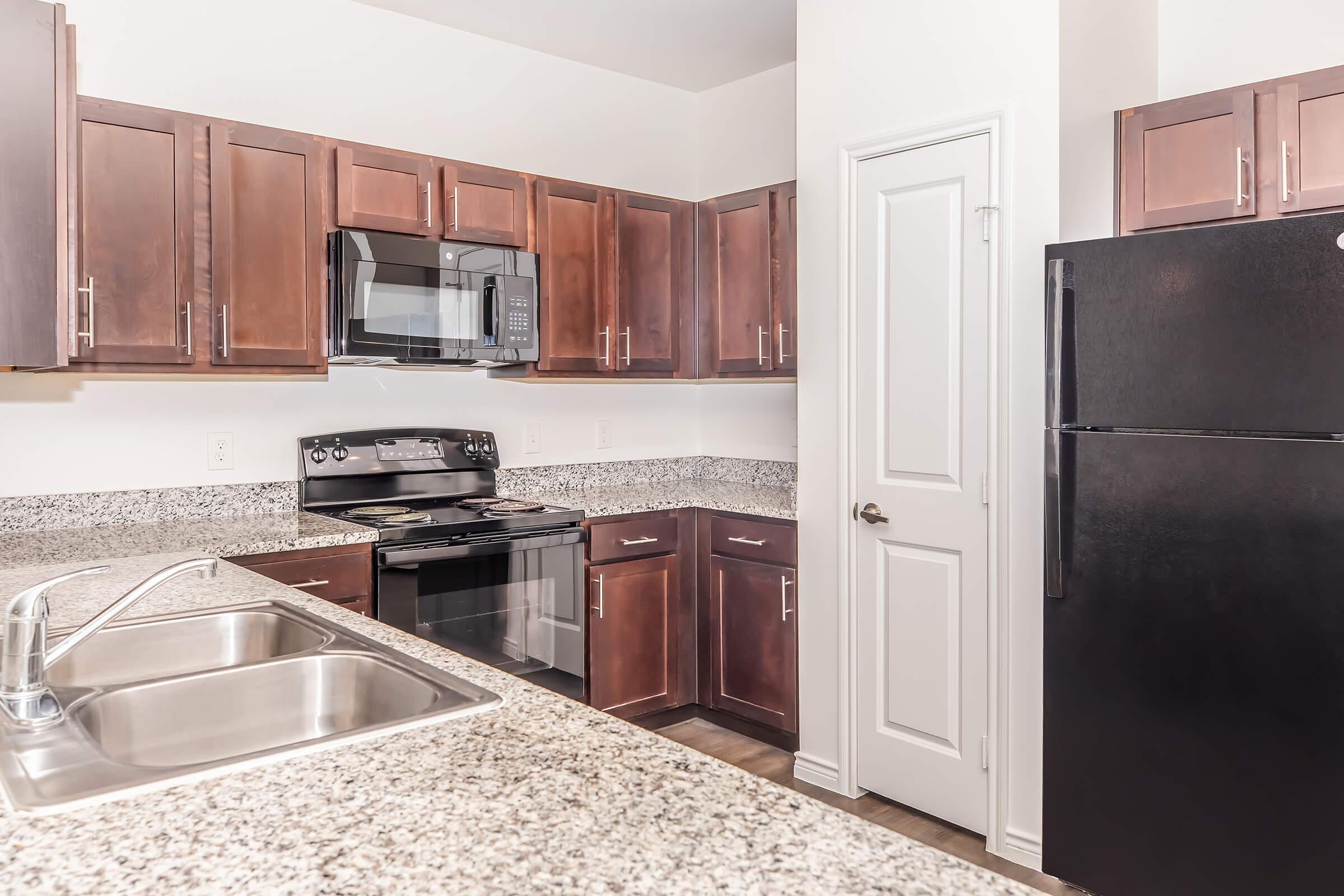 Modern kitchen featuring dark wood cabinets, stainless steel appliances, including a microwave and stove, a black refrigerator, and a granite countertop. The layout is open with a sink and ample cabinet space, providing a sleek and functional cooking area.