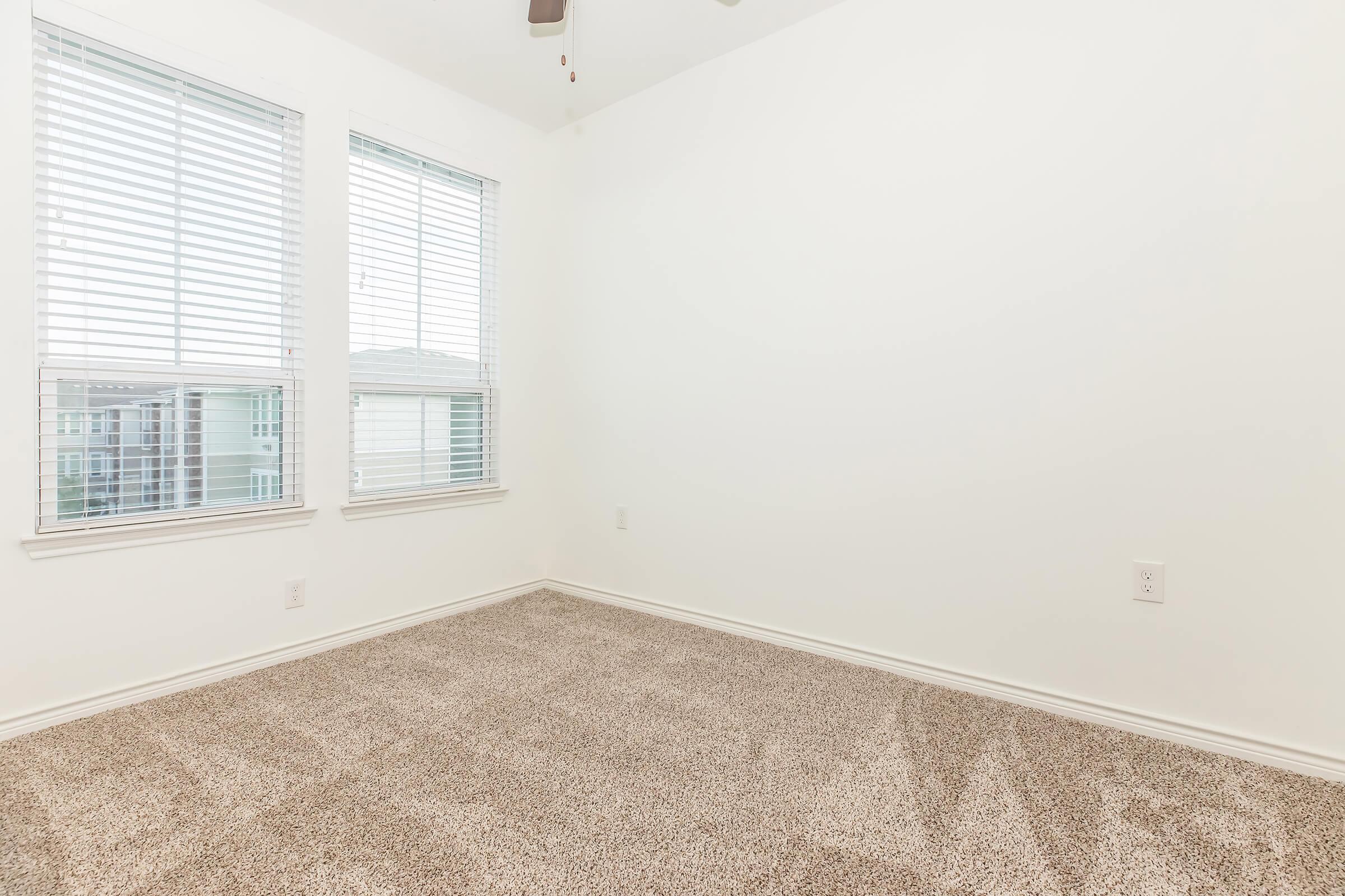 A vacant, empty room with light-colored walls and carpet. Two windows with blinds are positioned on the left side, allowing natural light to enter. The room features a ceiling fan and has a clean, minimalist aesthetic, making it suitable for various uses.