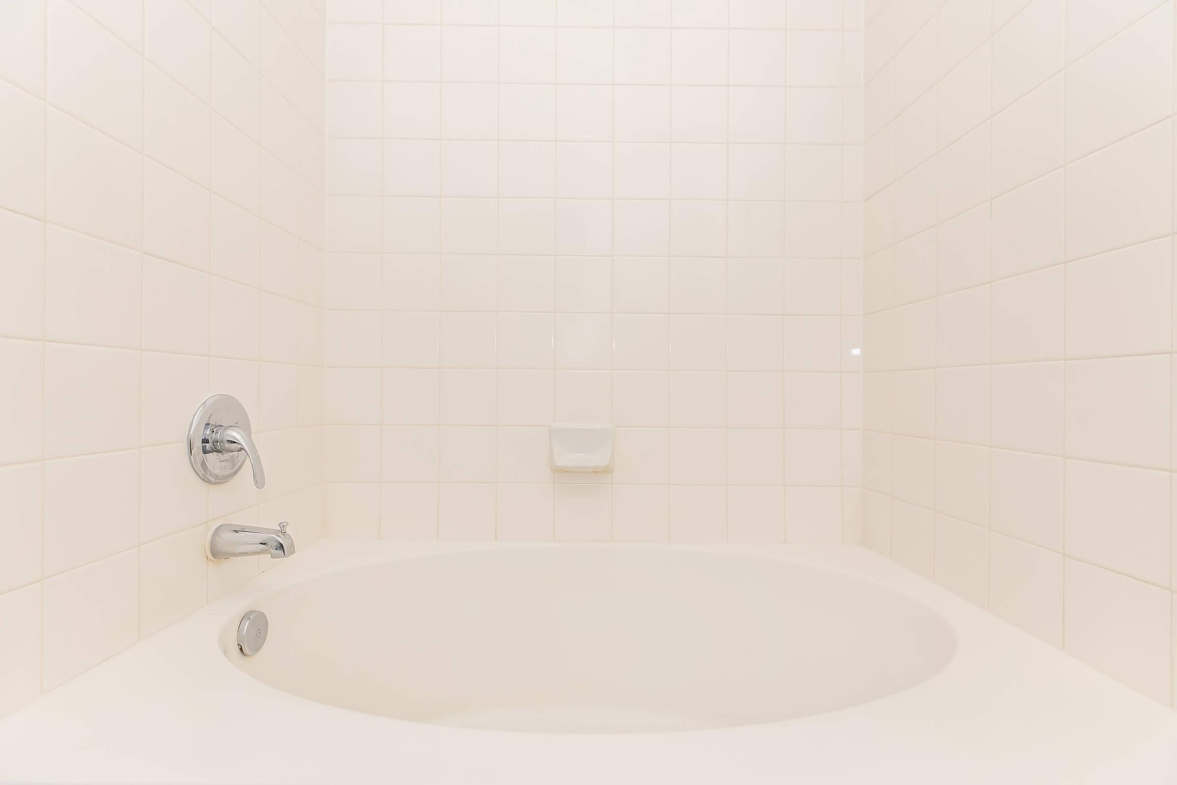 A clean, white bathroom featuring a round bathtub against a light-colored tiled wall. The faucet and spout are visible, with a soap dish mounted on the wall. The overall atmosphere is bright and minimalistic.