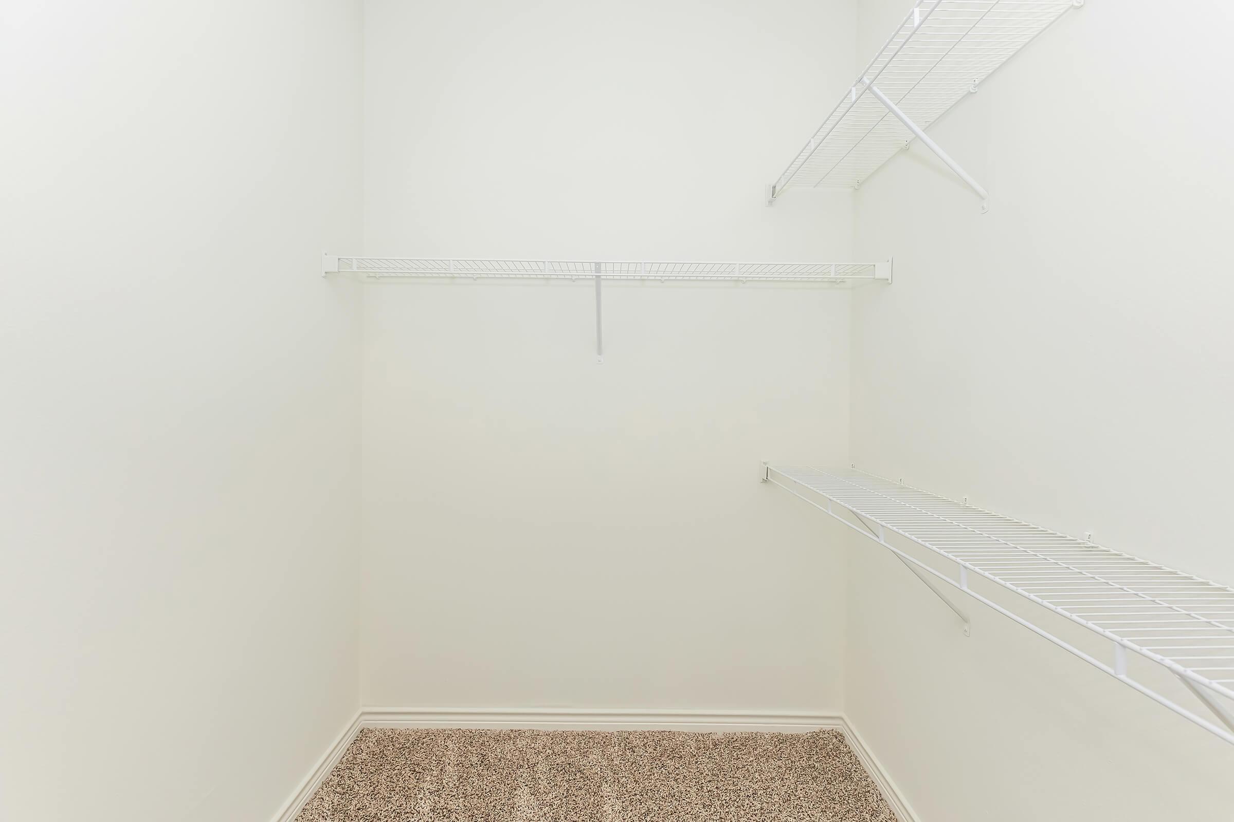 Empty closet interior with white wire shelving on two sides, light-colored walls, and a carpeted floor.