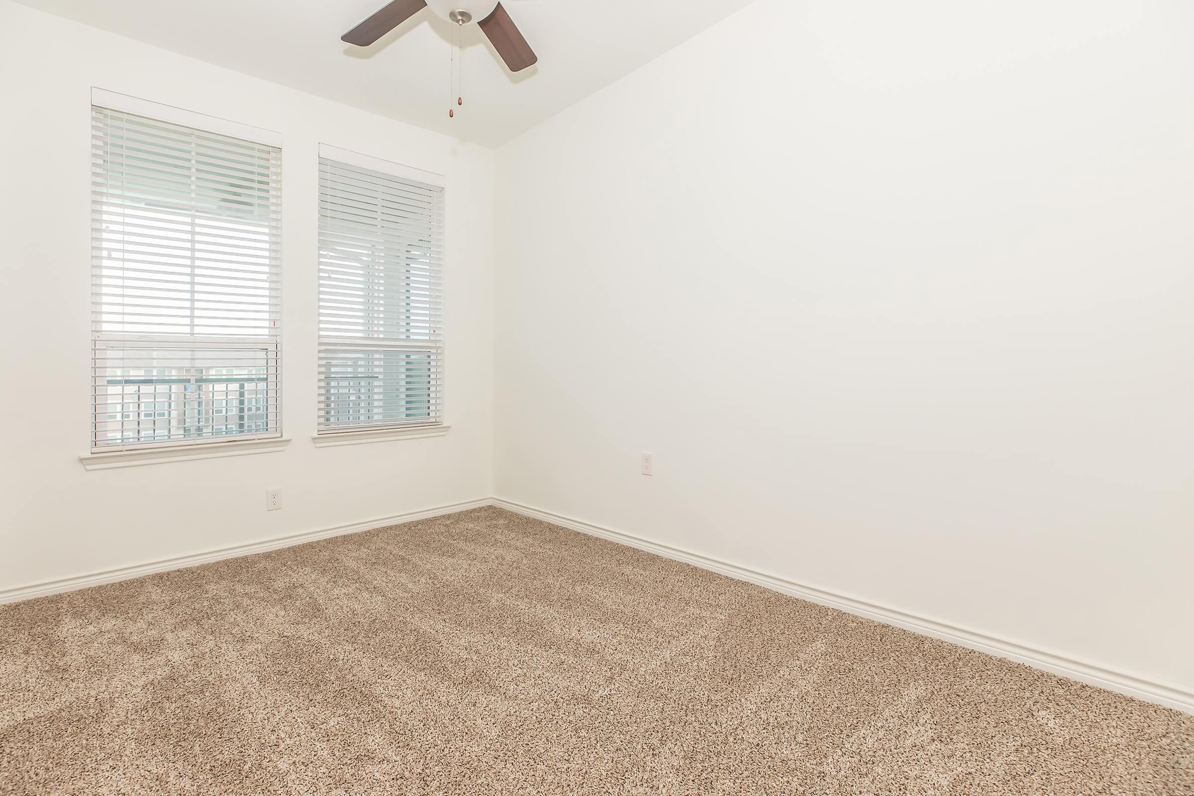A bright, empty room with beige carpet and white walls. There are two windows with blinds on the left side, providing natural light. A ceiling fan is visible in the center, and the overall atmosphere is clean and modern, suitable for personalizing as a living space.