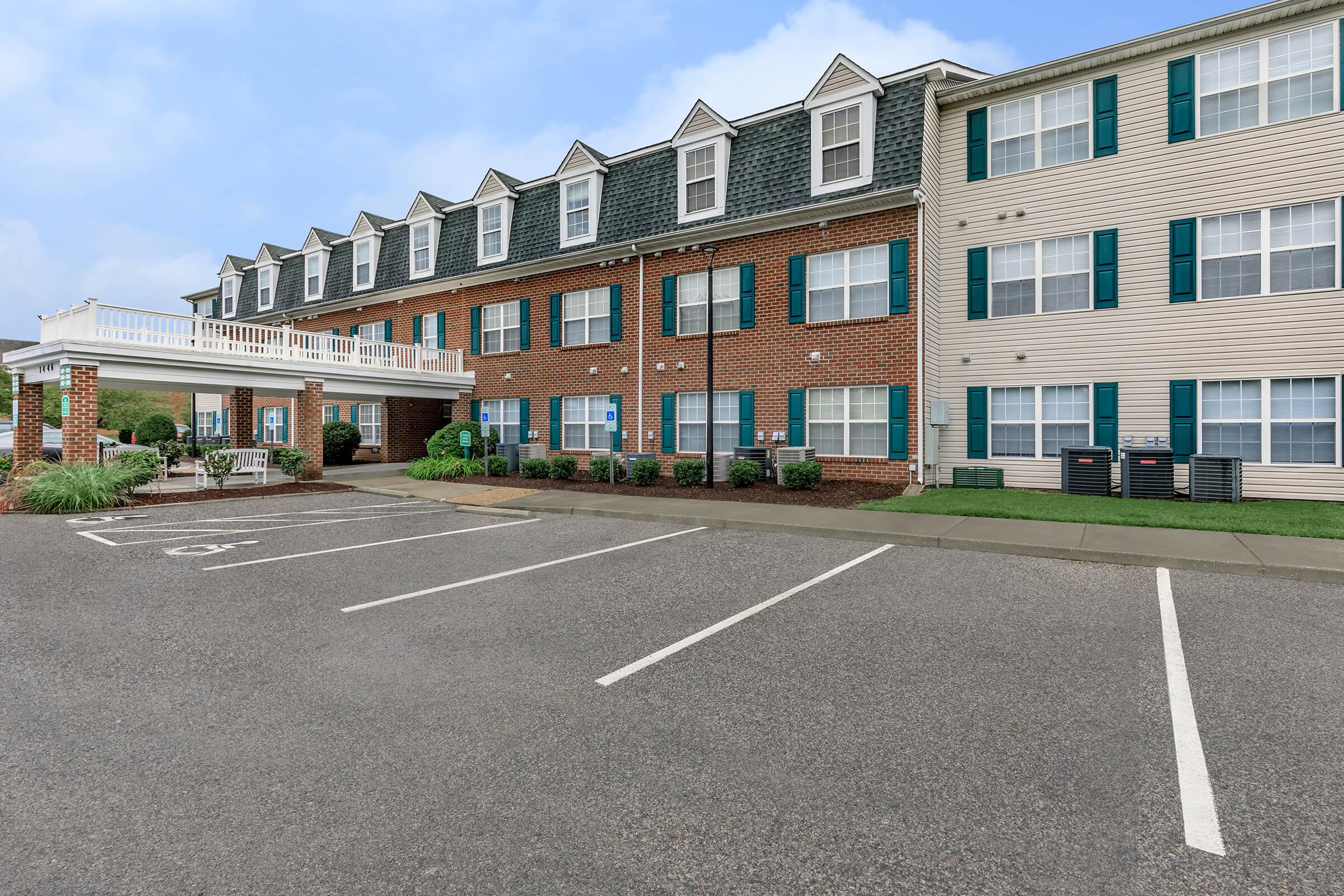 Two-story hotel exterior featuring brick and beige siding. The building has a sloped roof with dormer windows, green shutters, and a covered entrance. Parking spaces are visible in the foreground, with well-maintained landscaping alongside the pathway. The sky is partially cloudy.