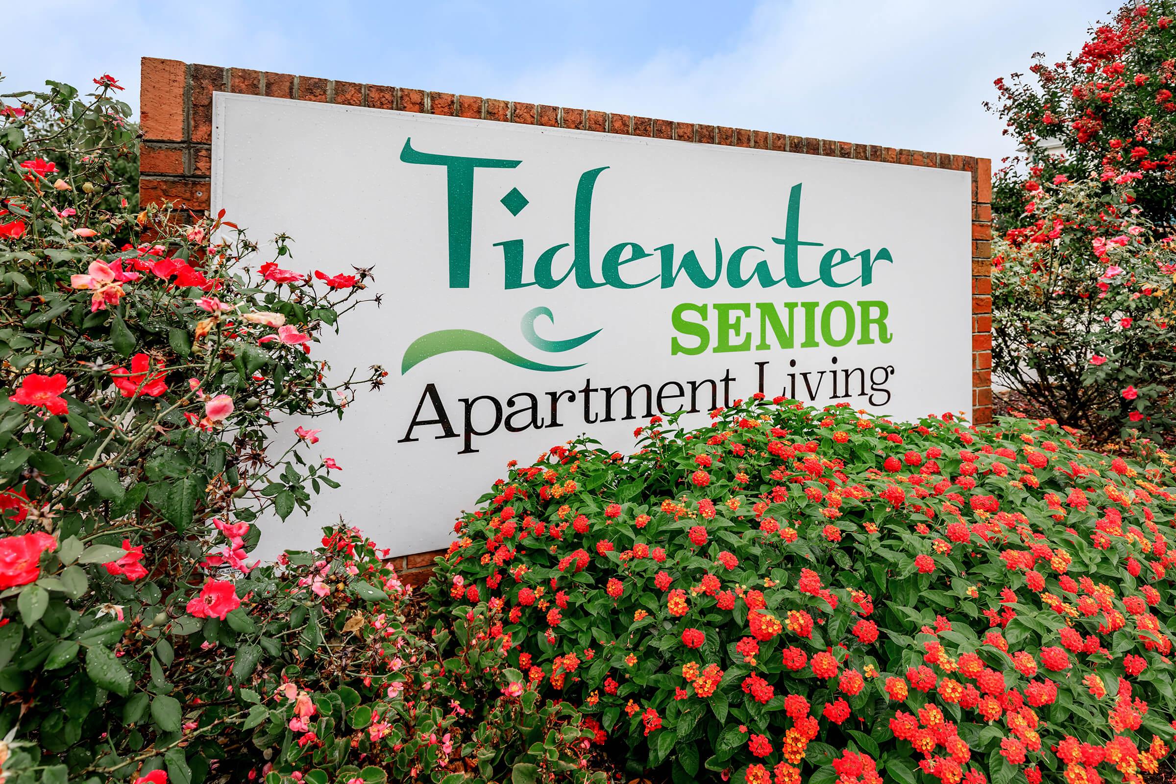 Sign for Tidewater Senior Apartment Living surrounded by colorful flowers. The sign features the name prominently, with decorative elements and greenery enhancing the inviting atmosphere of the senior living community.