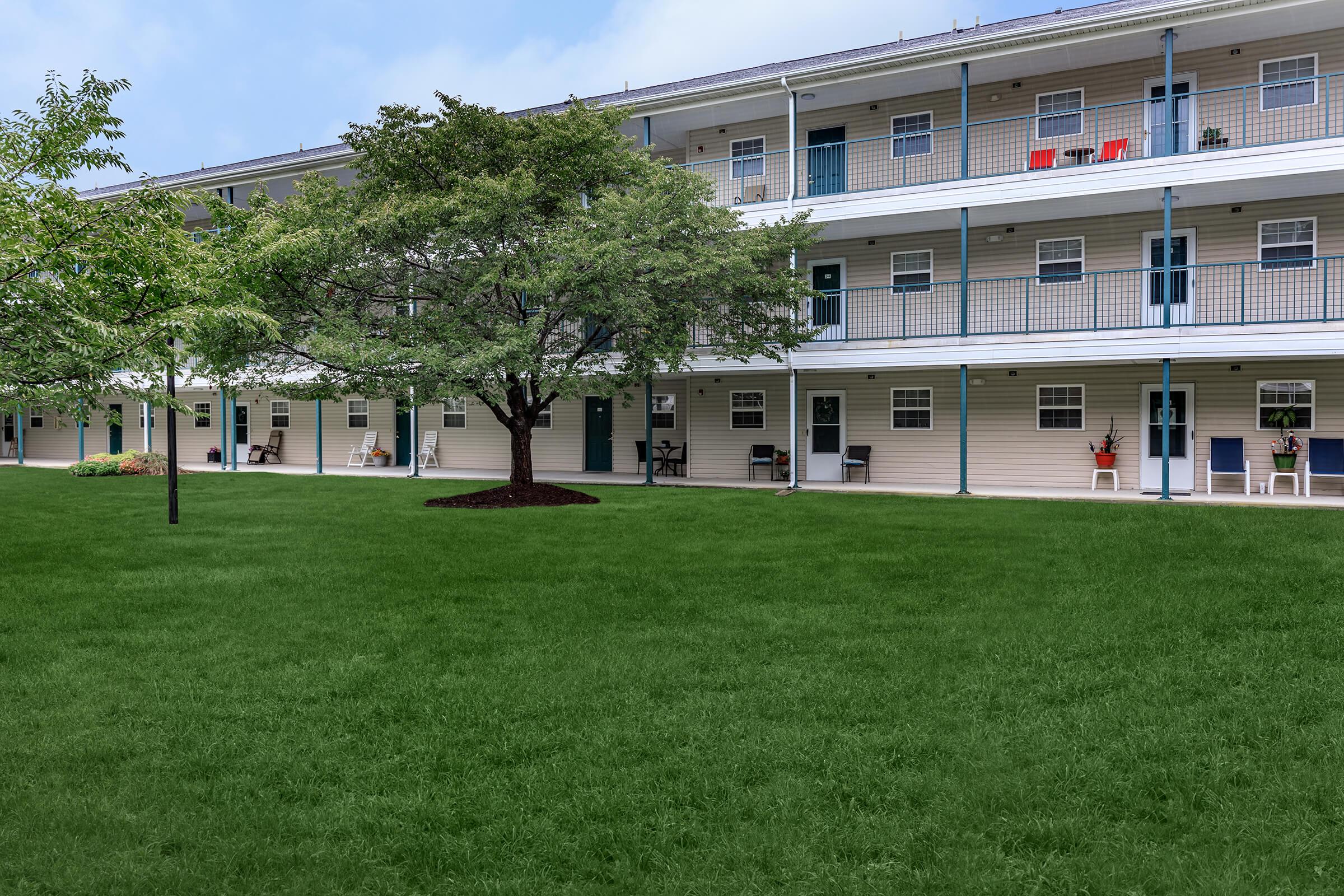 A well-maintained motel-style building with multiple balconies, featuring green grass in the foreground and trees, creating a pleasant outdoor space. Some balconies have chairs, and the overall setting appears clean and inviting under a clear blue sky.