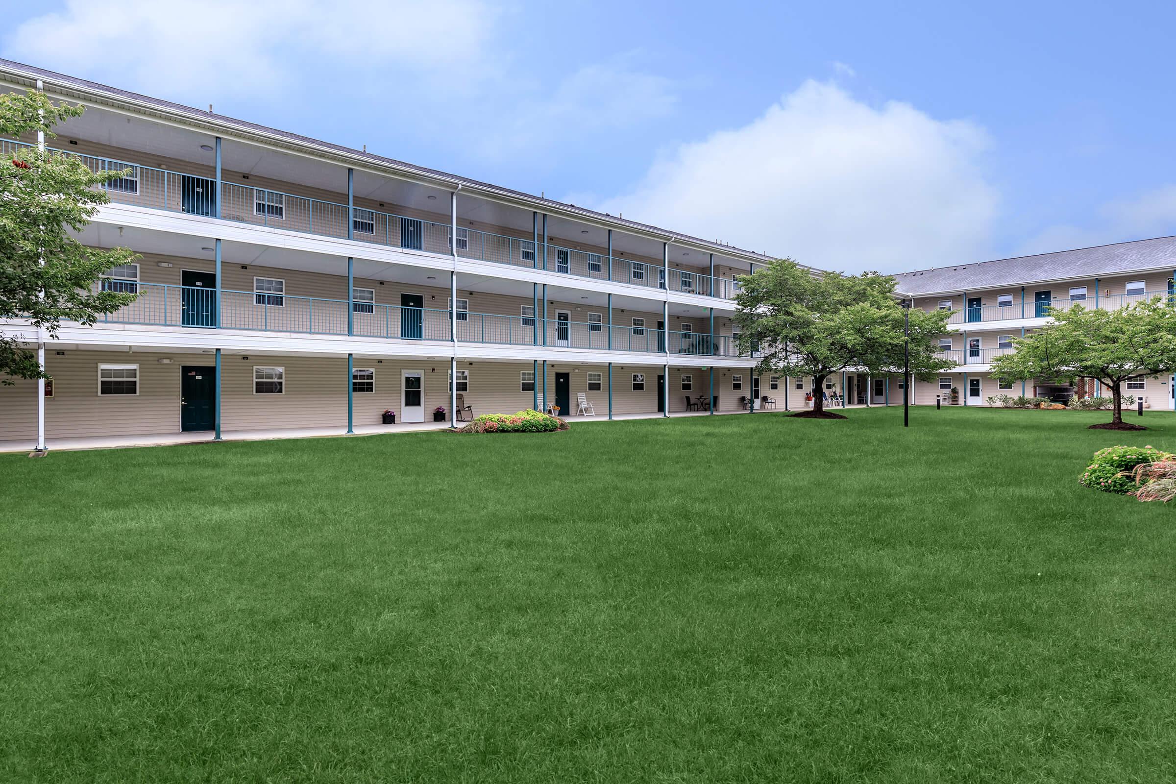 A view of a residential building with multiple stories, featuring balconies and green grassy areas in the foreground, surrounded by well-maintained trees and shrubs. The sky is clear with soft clouds. The setting suggests a calm and inviting apartment complex.