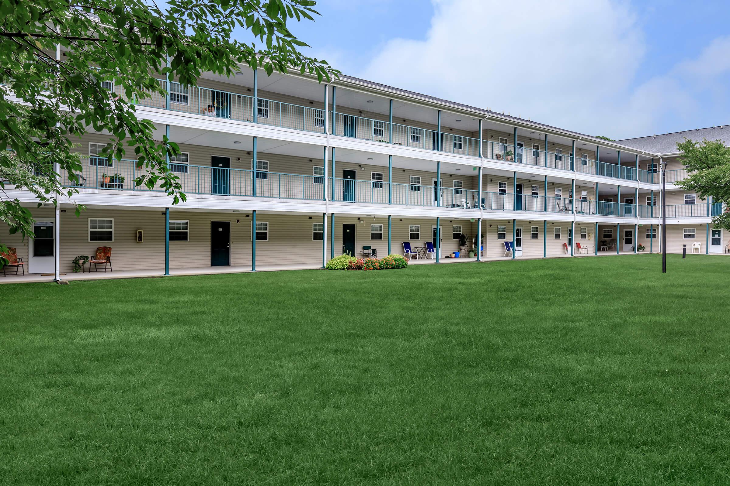 Three-story apartment building with multiple balconies and doors on each floor. Lush green lawn in front, with a few chairs placed on the balconies. Overcast sky in the background.
