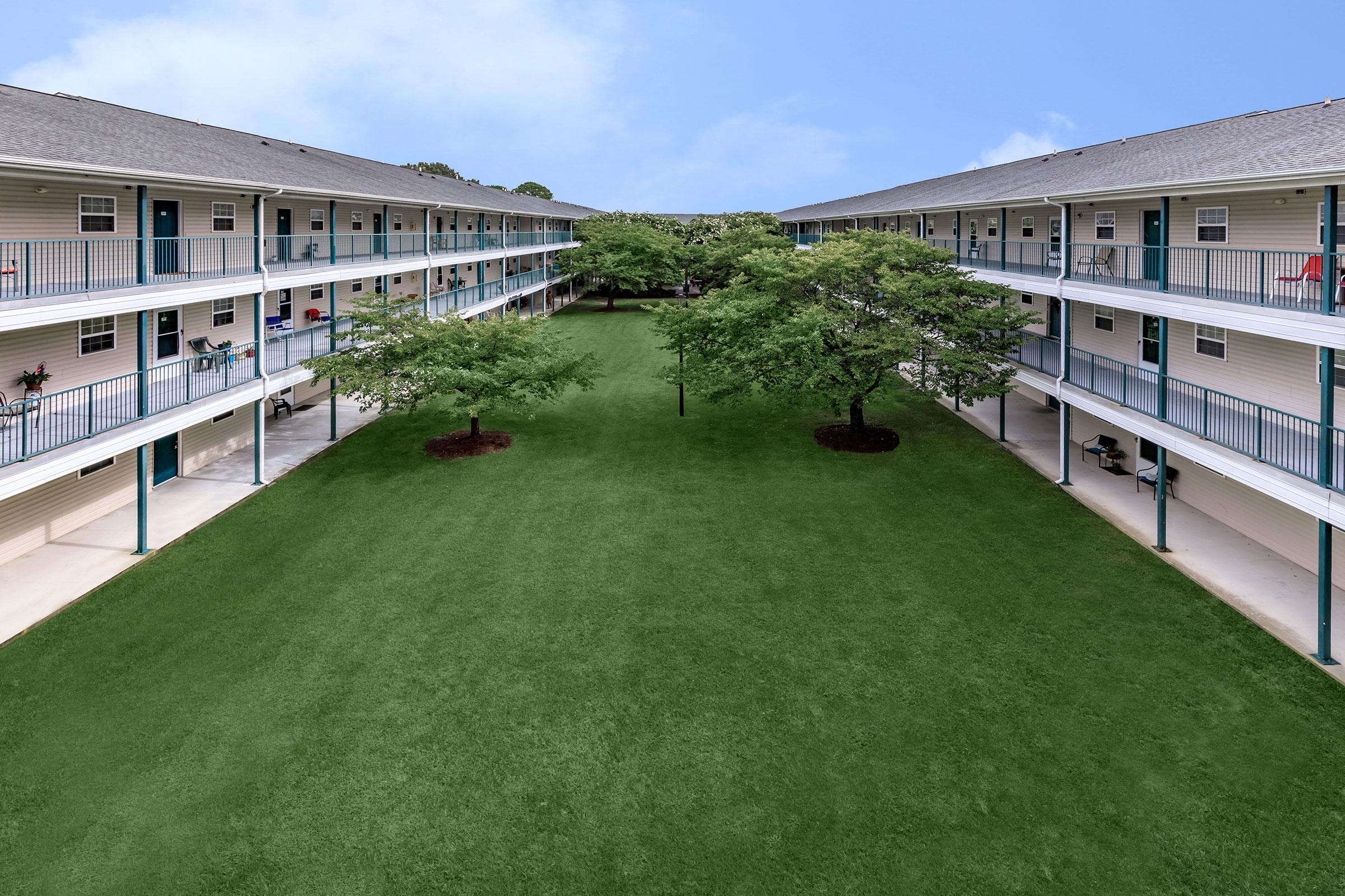 Aerial view of a two-story apartment complex with two rows of units facing a grassy courtyard. The courtyard features two small trees, and the surrounding balconies are lined with railings. The sky is clear and blue, enhancing the appearance of the buildings.