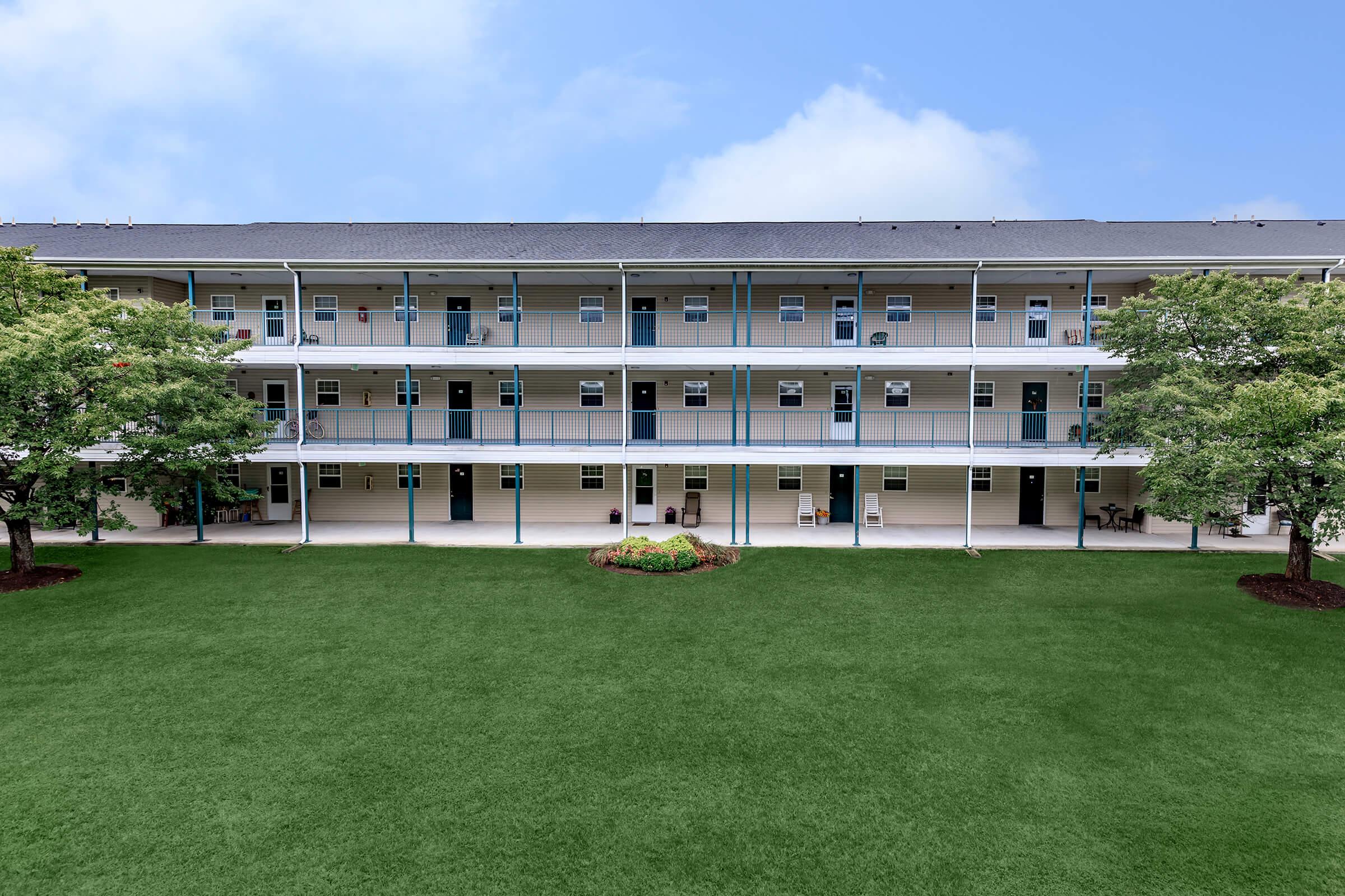 A three-story residential building with multiple balconies and light-colored walls, surrounded by a well-maintained green lawn and a small landscaped area with flowers. The sky is clear and bright, enhancing the overall pleasant atmosphere of the location.