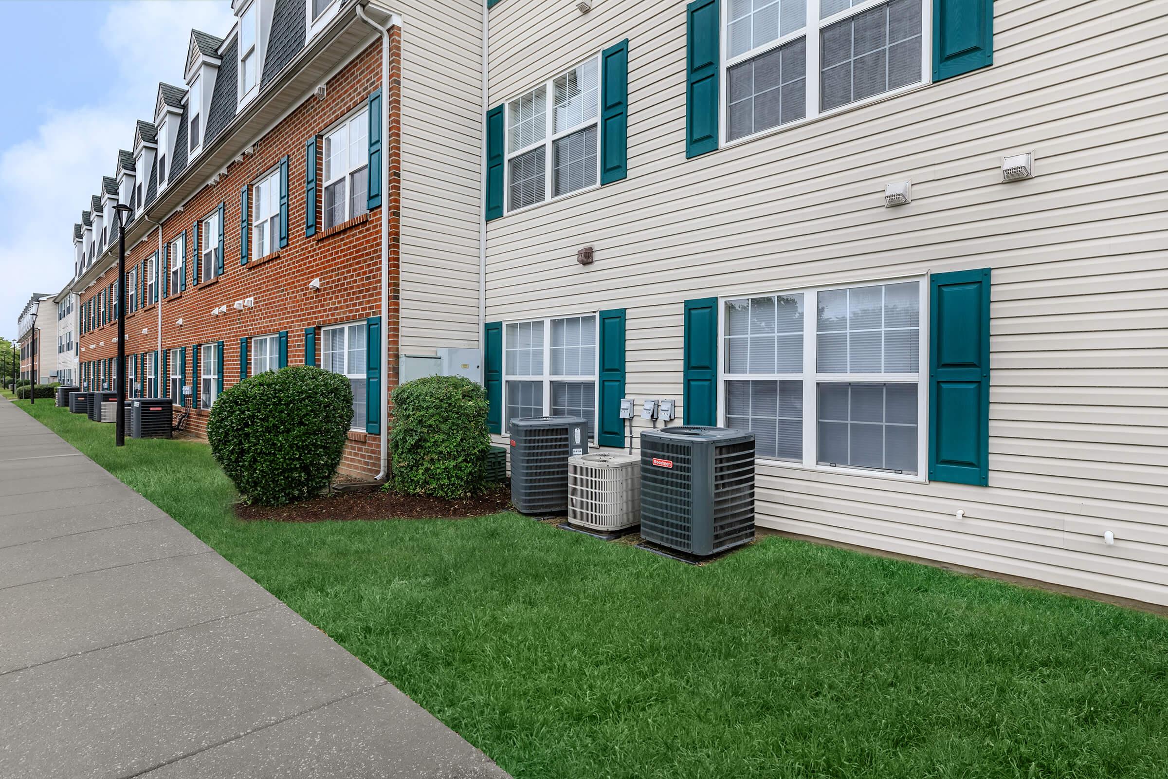 A view of a residential building exterior featuring a sidewalk, green lawn, and several air conditioning units beside the building. The structure includes both brick and siding, with multiple windows and decorative shutters in teal color.
