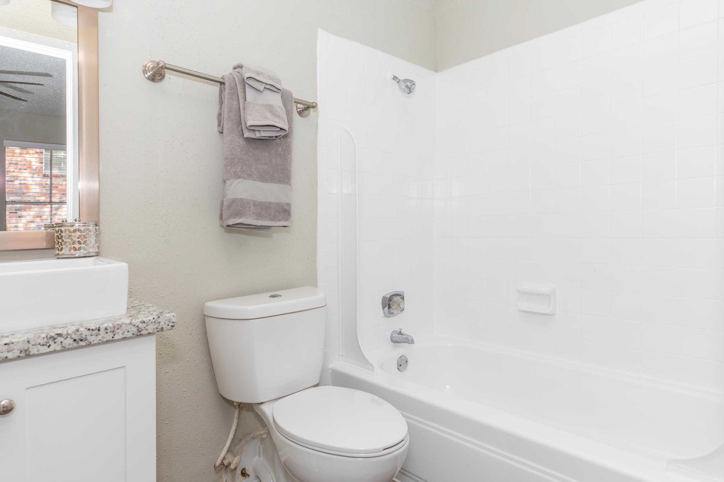 A clean, modern bathroom featuring a white bathtub with shower fixtures, a toilet, and a small sink with a granite countertop. A towel is hanging on a bar beside the bathtub, and the walls are painted a light color, enhancing the overall brightness of the space.