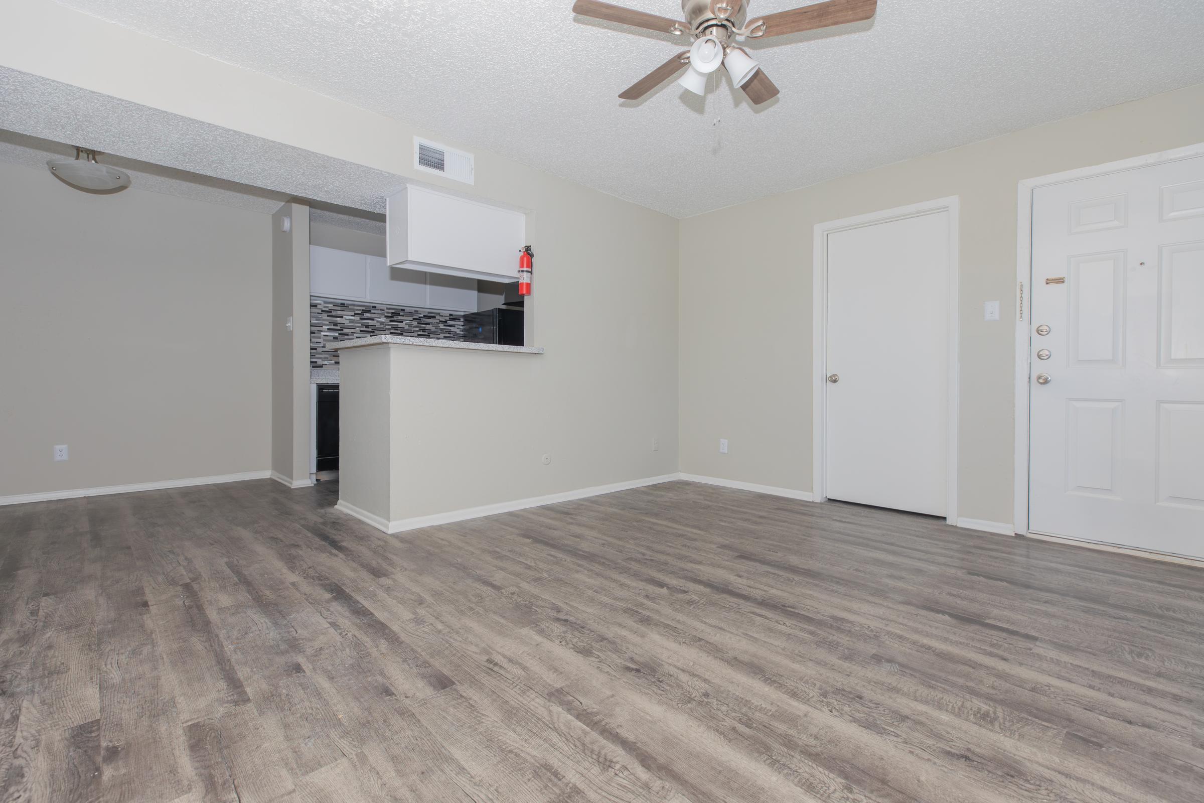 A spacious interior of an apartment featuring light-colored walls, a ceiling fan, and laminate flooring. A doorway leads to a kitchen area with modern design. The front door is visible, and there is a small dining area in the background. Natural light brightens the room, creating an inviting atmosphere.