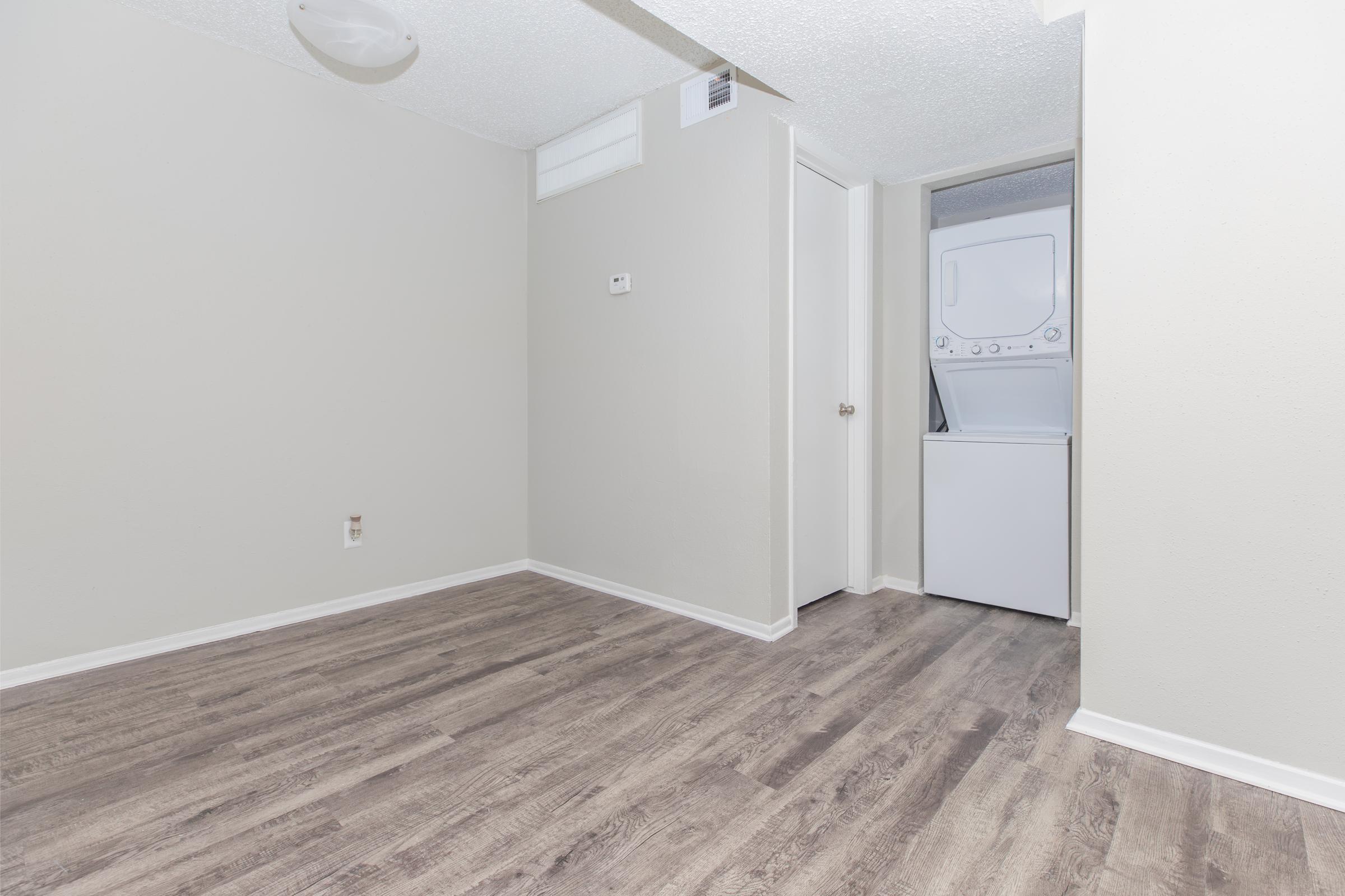 A spacious, empty room with light gray walls and new laminate flooring. There is a small wall vent and a door leading to a laundry area featuring a washer and dryer. The room is well-lit, giving it a clean and modern look.