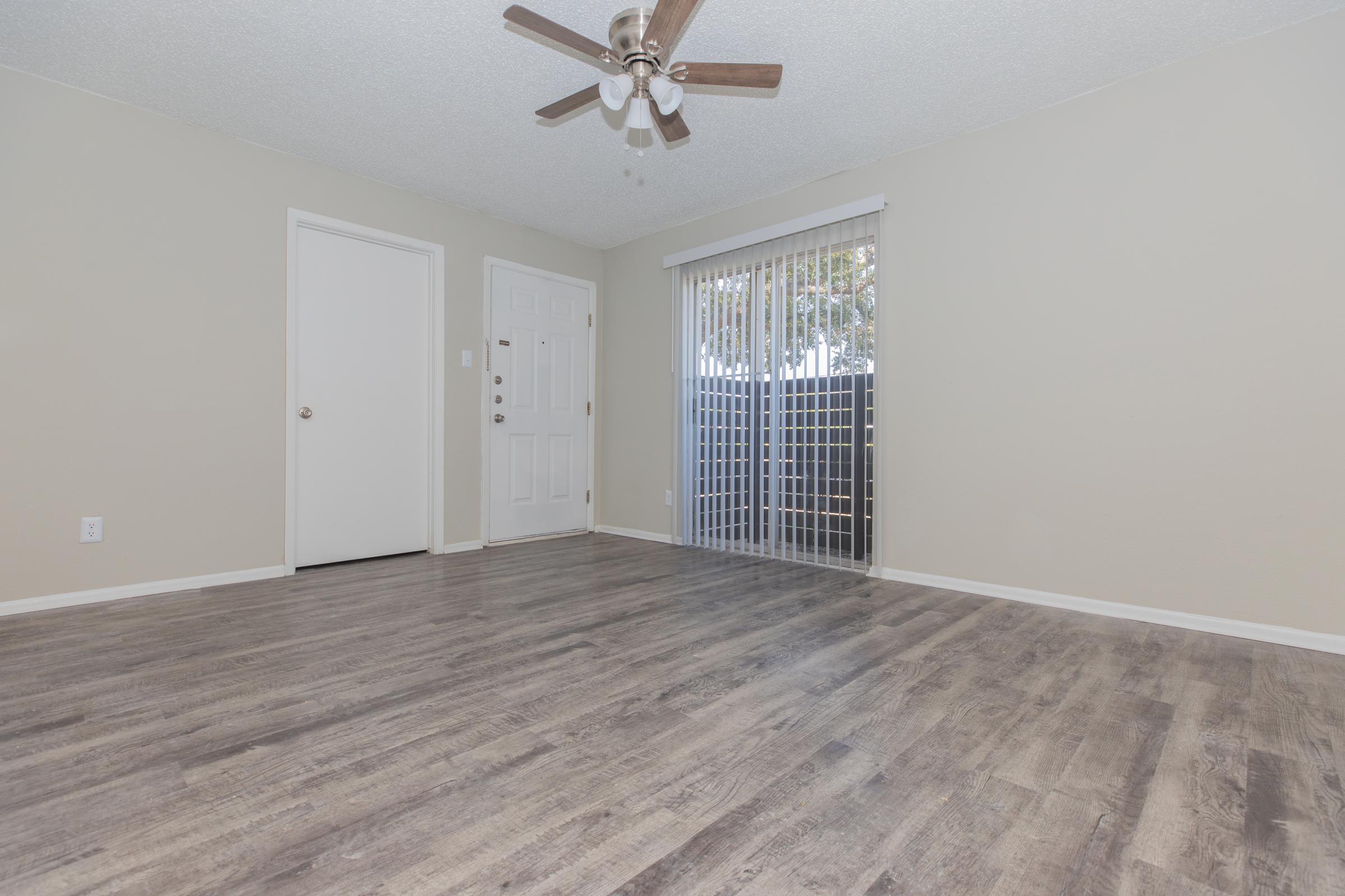 A spacious, empty room with light-colored walls and a ceiling fan. There's a white door on the left, leading to another room, and a large window covered with vertical blinds that allows natural light to enter. The floor is a light oak laminate, creating a warm and inviting atmosphere.
