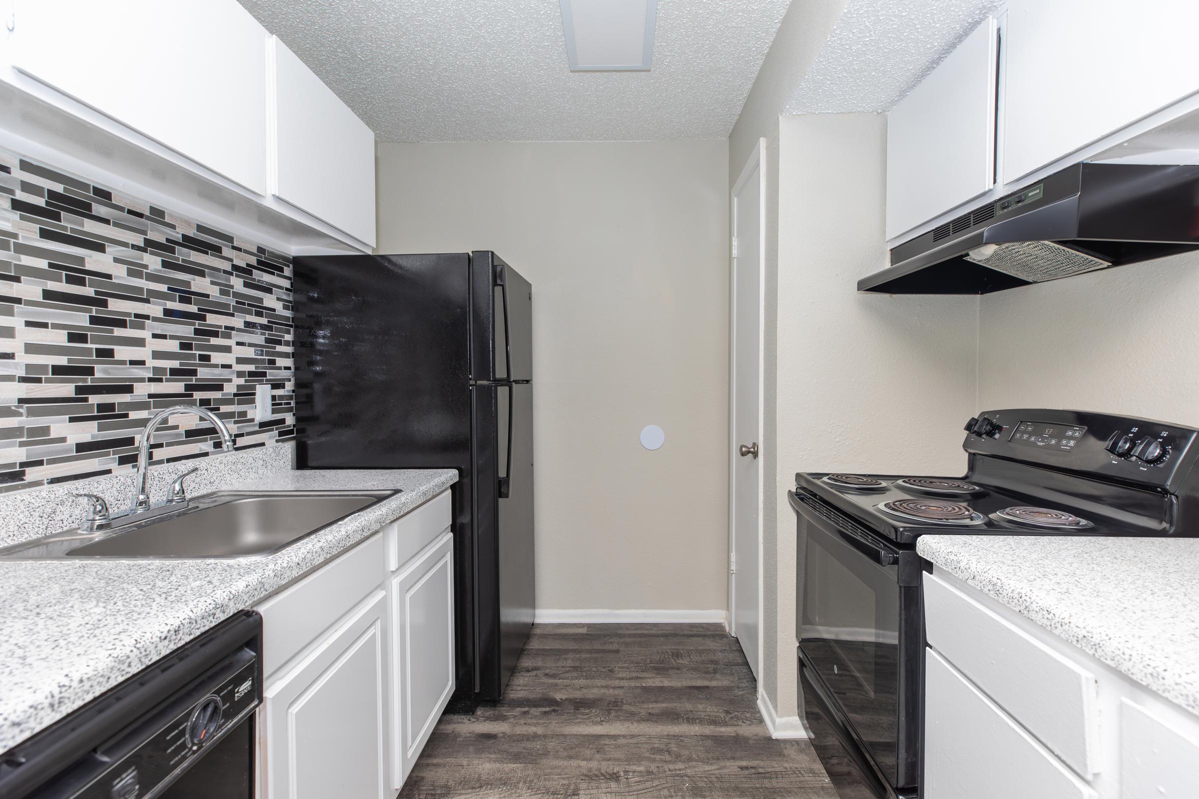A modern kitchen featuring black appliances, including a refrigerator and an oven. The countertops are a light granite-like material, complemented by a stylish backsplash of black and gray tiles. The cabinets are white, and the flooring is dark laminate, creating a contemporary look.
