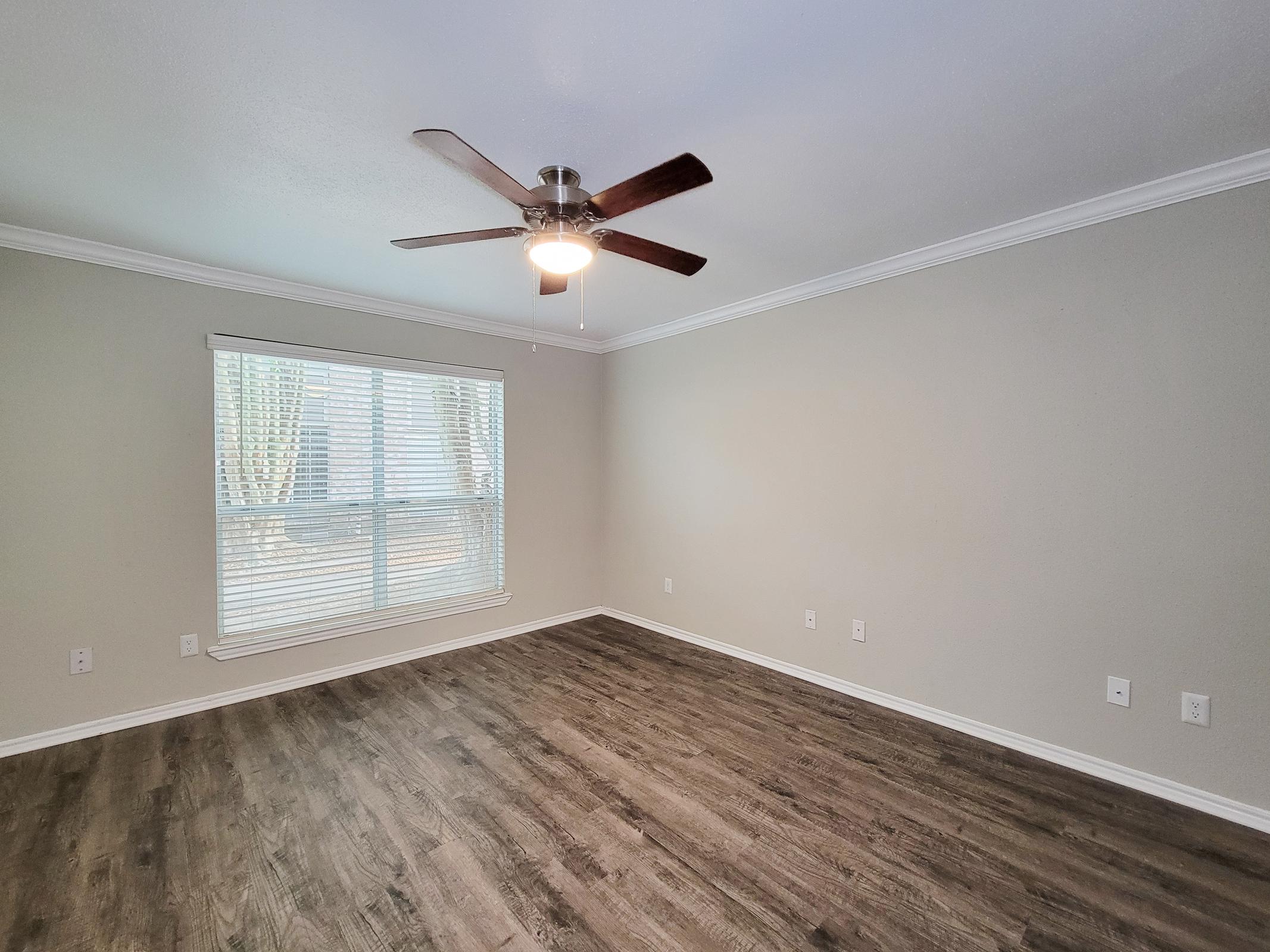 Spacious, empty living room featuring a ceiling fan, large window with blinds, and light-colored walls. The flooring is a modern wood-like laminate, creating a warm atmosphere. Natural light floods the space, highlighting the openness and potential for personalization.