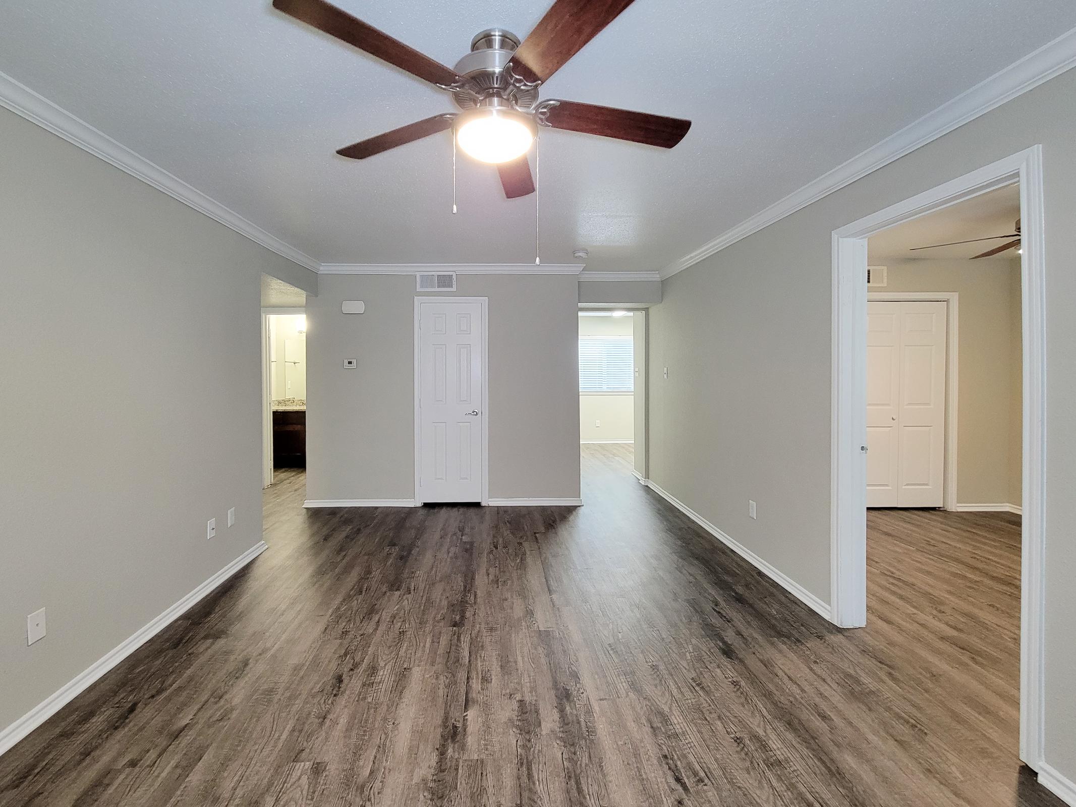 A spacious living area featuring light-colored walls and dark laminate flooring. A ceiling fan hangs from the center ceiling, providing ventilation. Two doorways lead to other rooms, and natural light is visible through a window in the background. The overall atmosphere is bright and inviting.